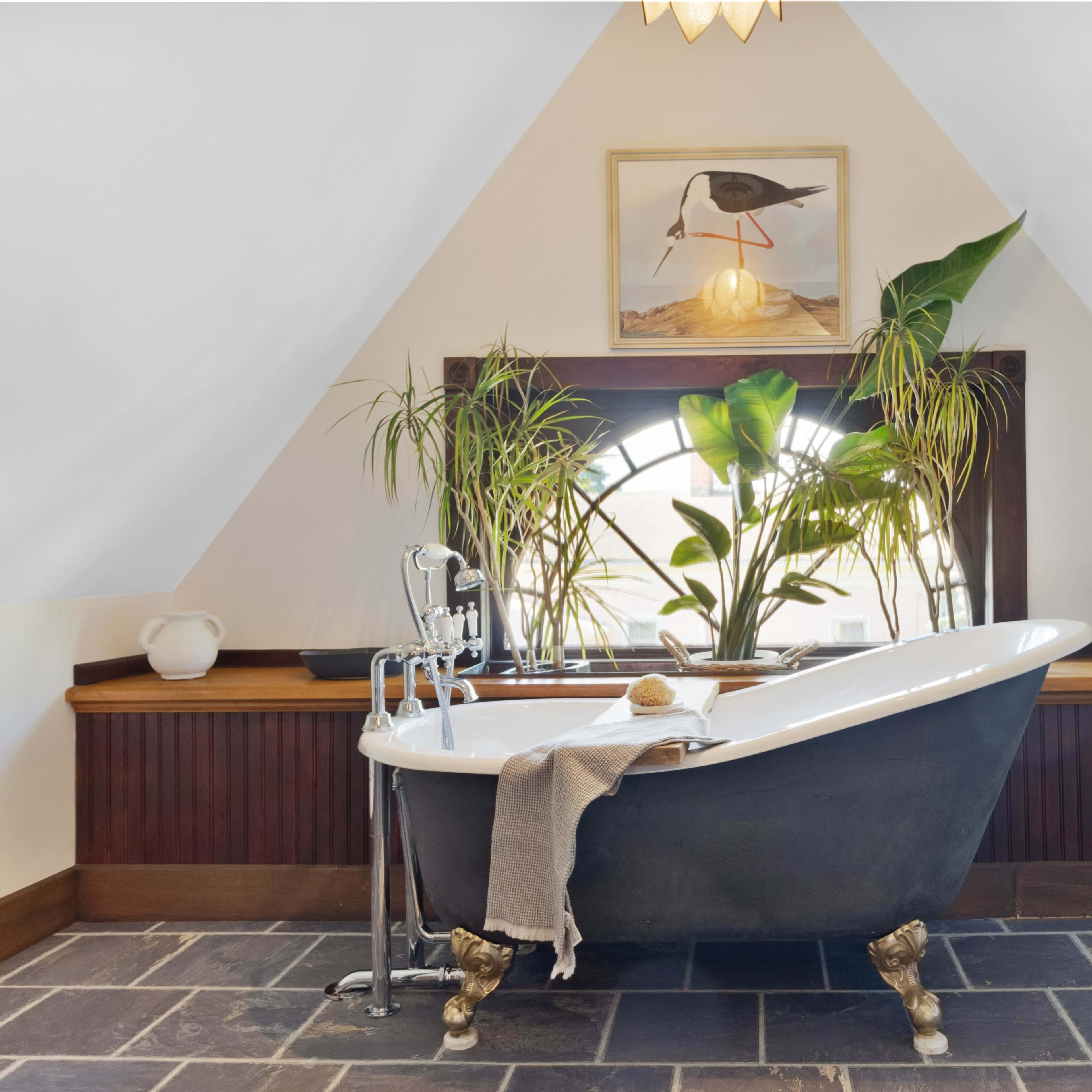 A freestanding bathtub is positioned near a large window surrounded by plants in a well-lit, triangular-shaped bathroom.