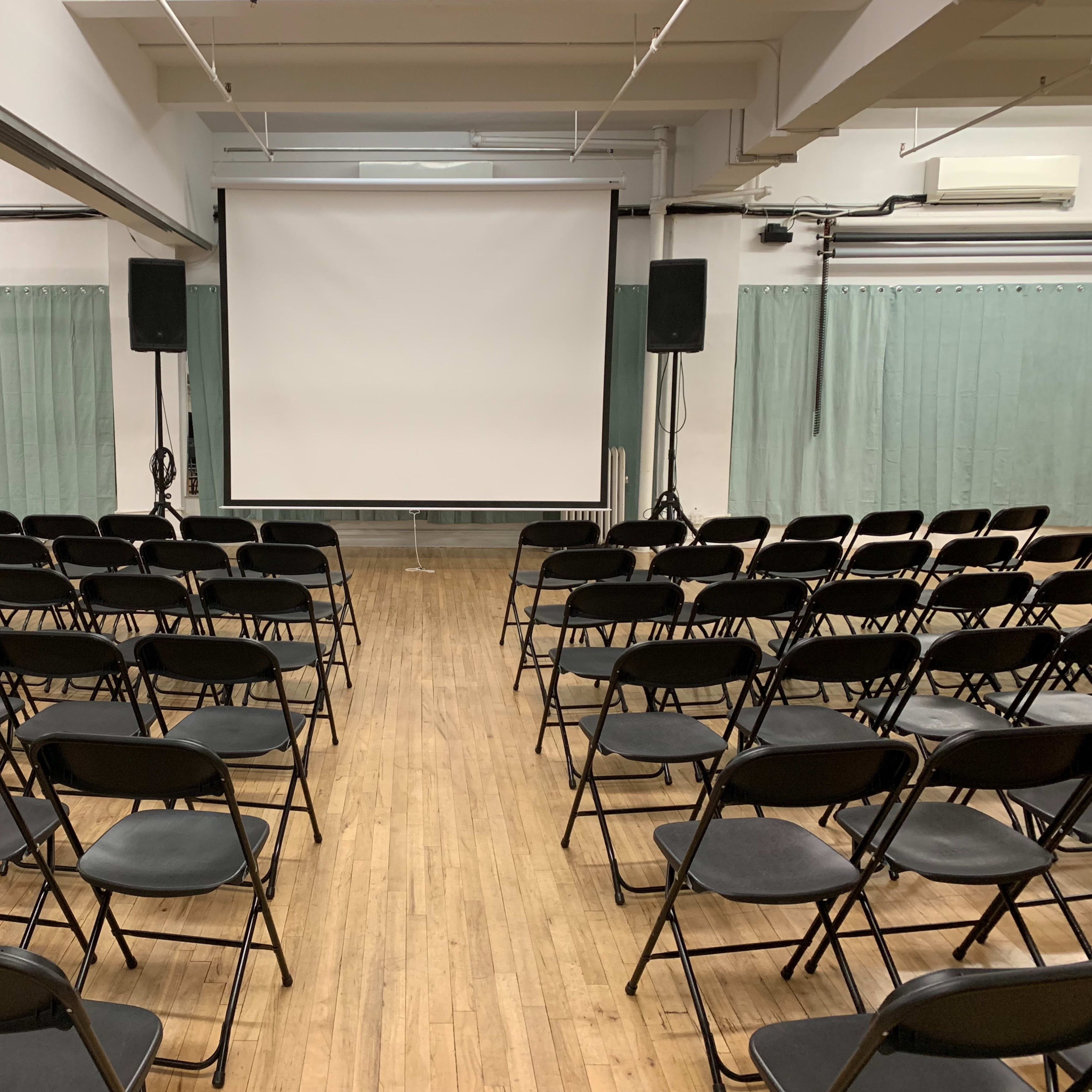 A large, mostly empty room with black folding chairs arranged in rows facing a blank projection screen.
