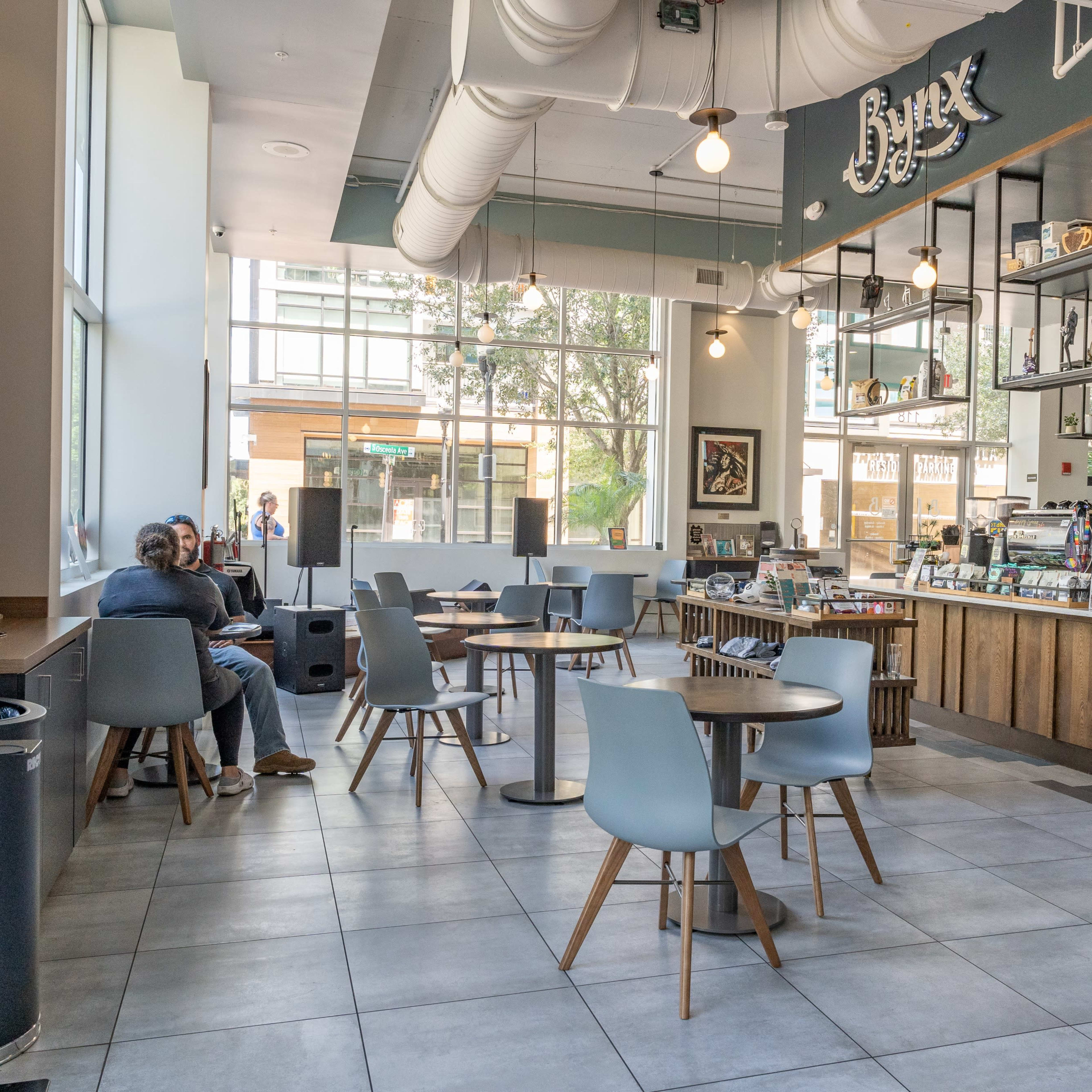A modern café interior with several gray chairs and tables arranged around a spacious area, featuring a counter with various items displayed and large windows letting in natural light.