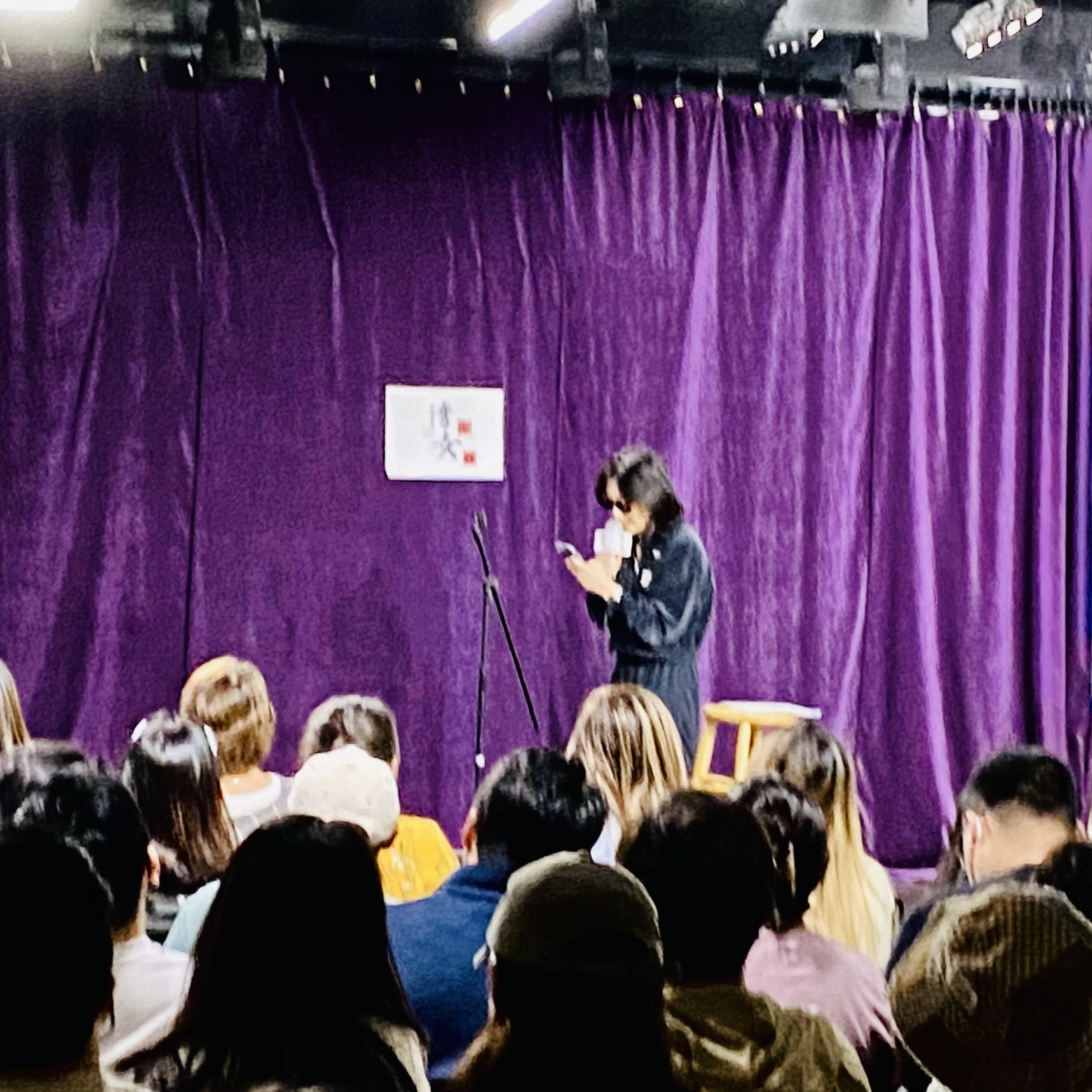 A performer stands near a microphone on stage while an audience sits in front, facing a purple backdrop.