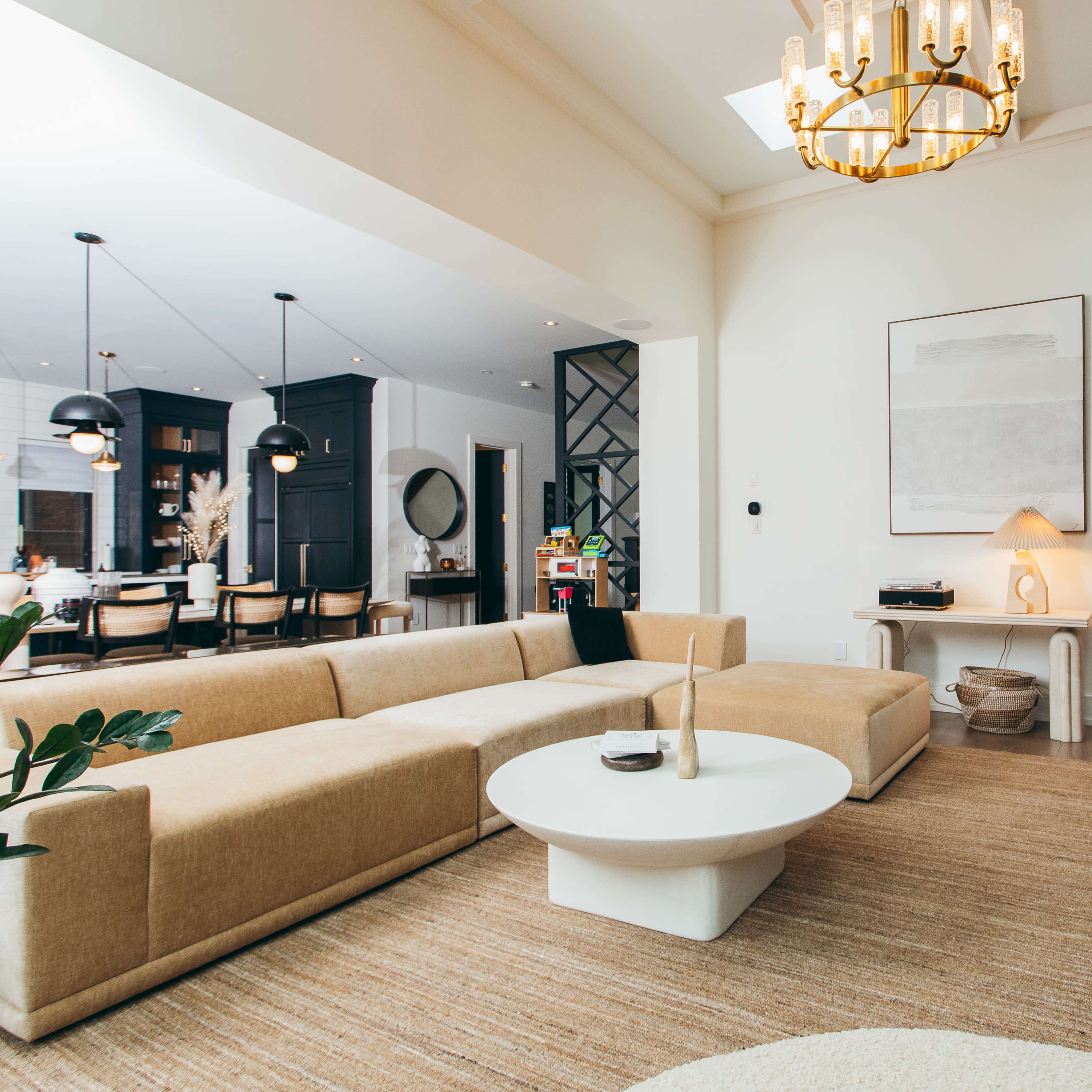 A modern living room with a large beige sectional sofa, a round white coffee table, and light-colored furniture, complemented by a chandelier and a minimalistic artwork on the wall.