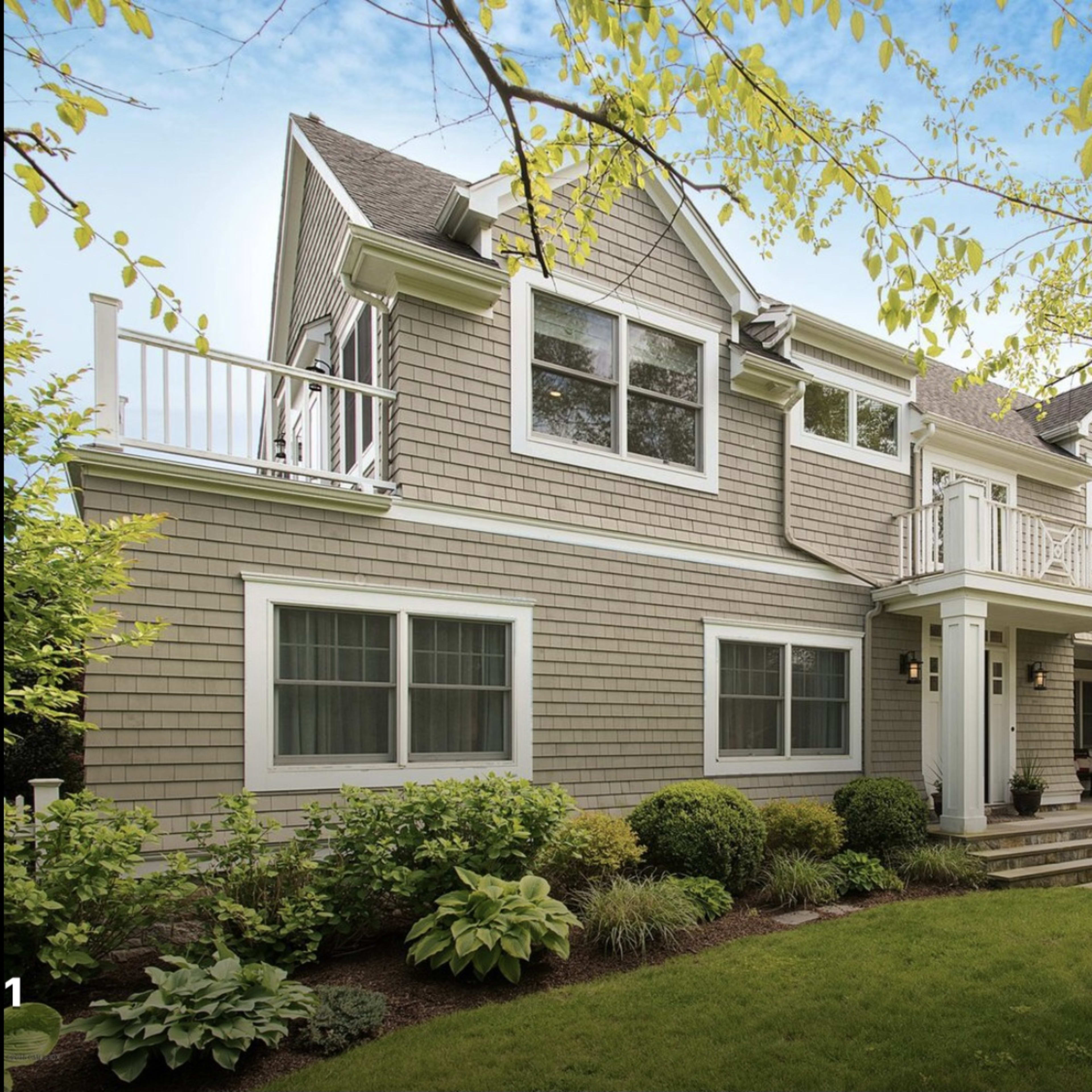 A two-story house with a light gray facade and a front porch is surrounded by greenery and small bushes.