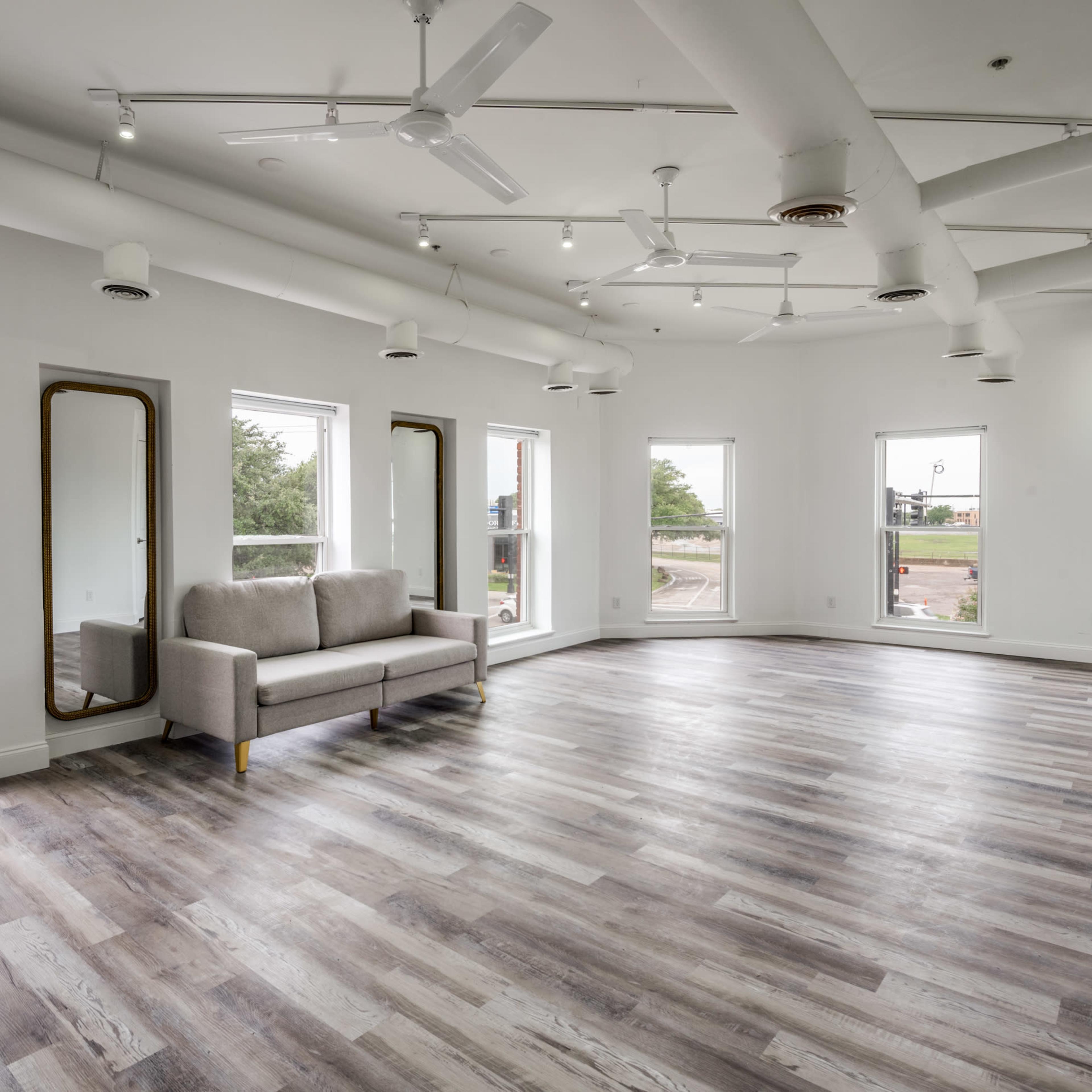 A spacious, well-lit room with hardwood flooring, two large windows, a gray couch, and mirrors on the walls.