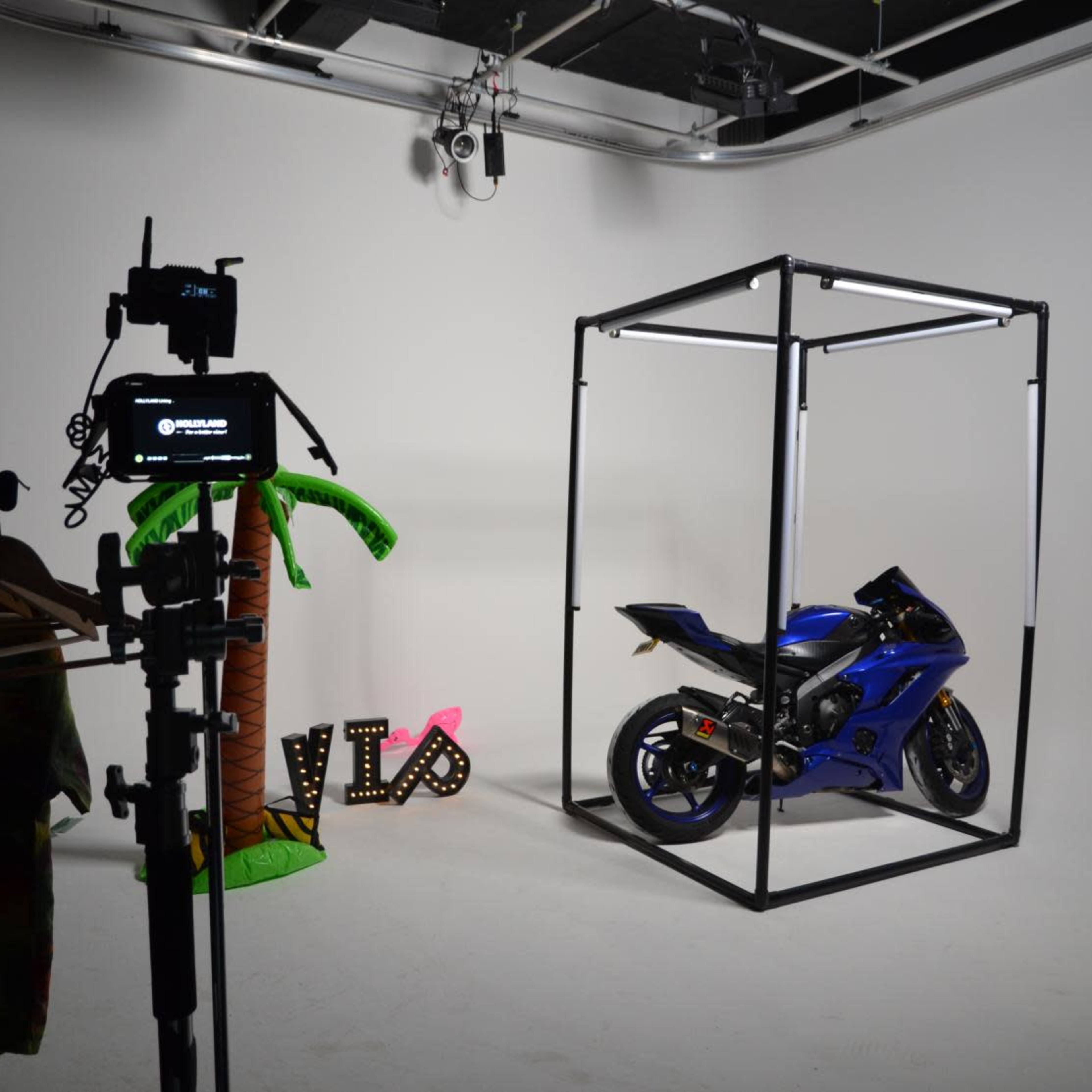 A blue motorcycle is displayed inside a metal frame on a plain background, with a palm tree decoration and a "VIP" sign nearby.