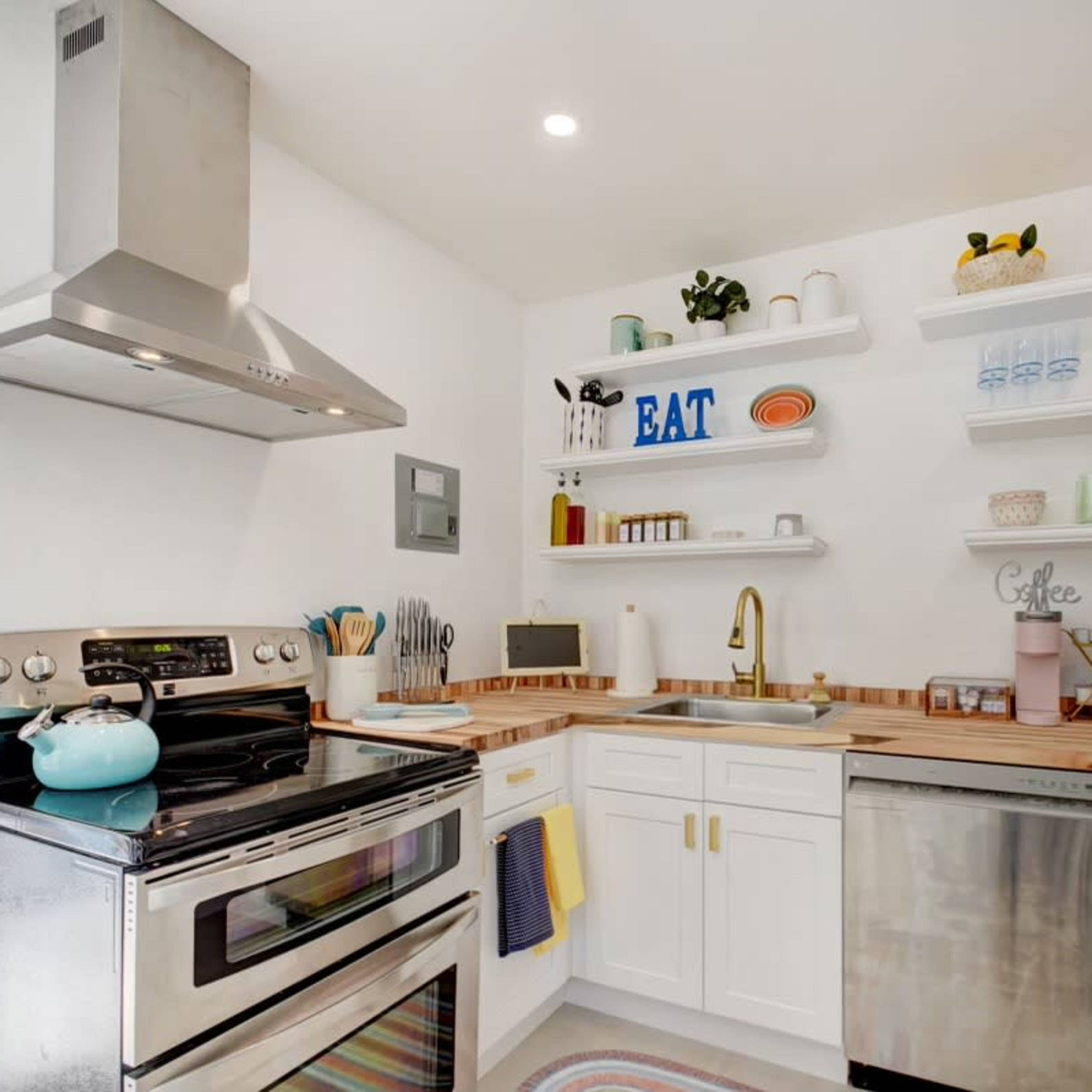 A modern kitchen with white cabinets, a stainless steel stove, and open shelves displaying colorful dishes and decor.