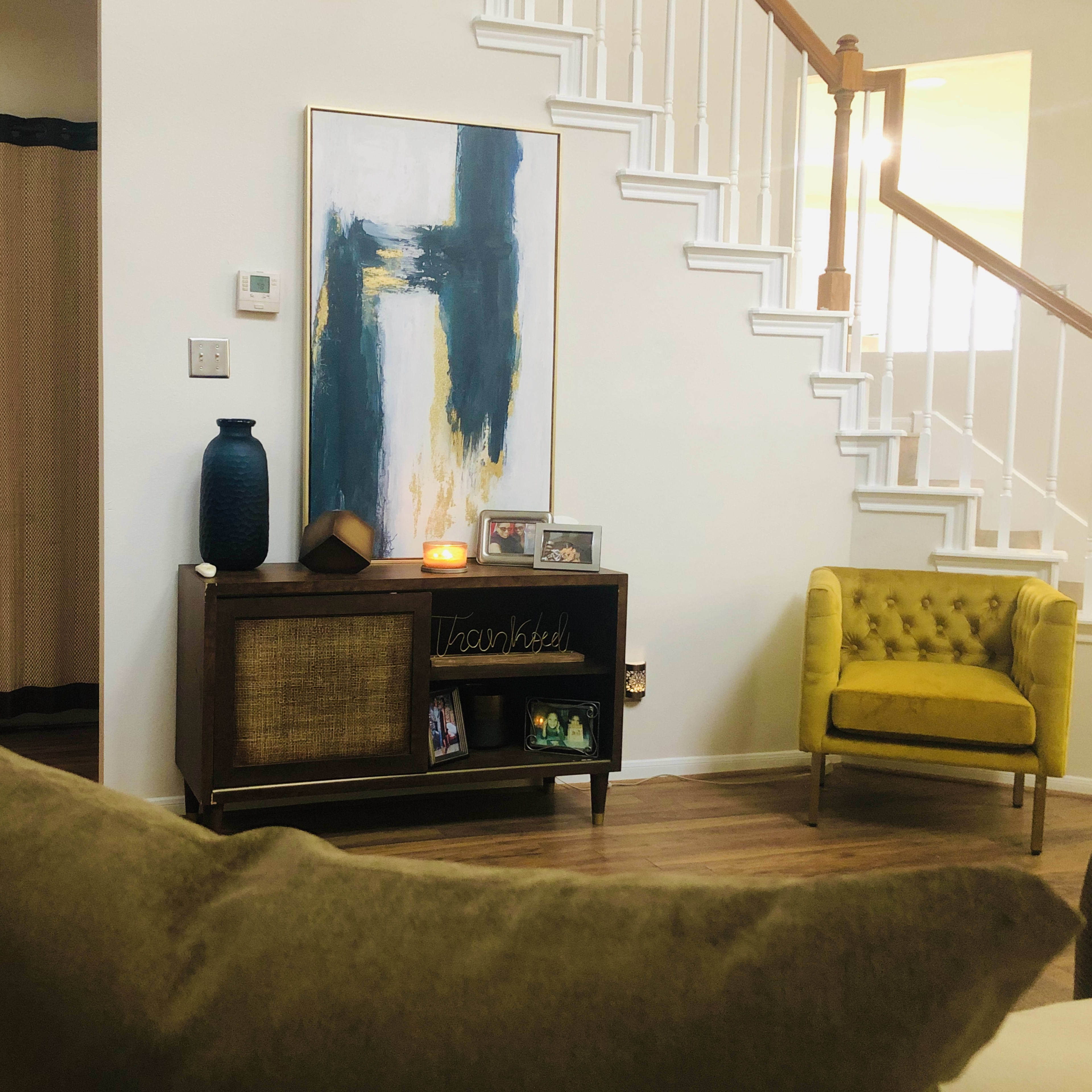 A cozy living room featuring a green sofa, a yellow armchair, a wooden console table with decor, and a large abstract painting on the wall.