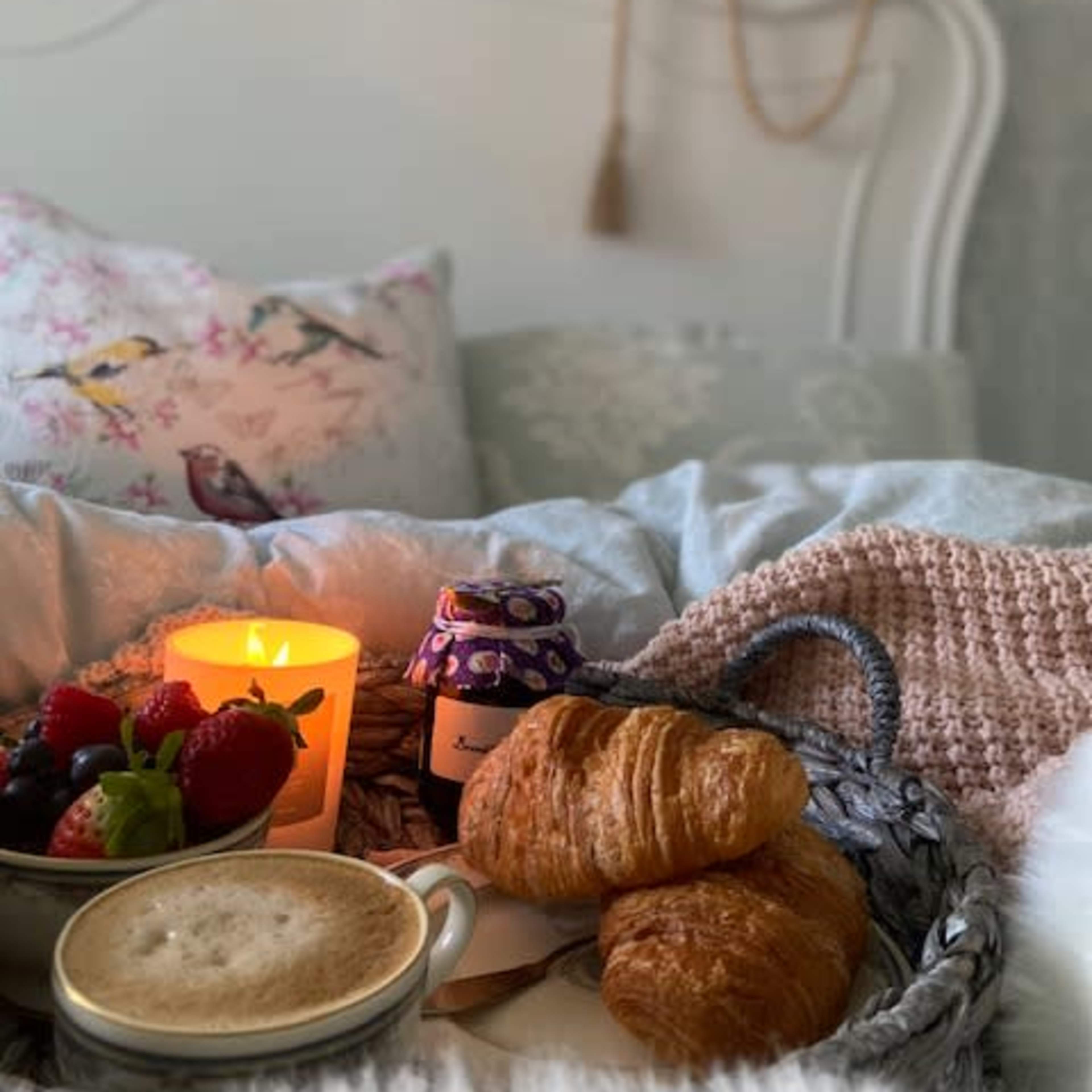 A cozy breakfast tray rests on a bed, featuring a cup of coffee, croissants, fresh berries, a candle, and a jar of jam.