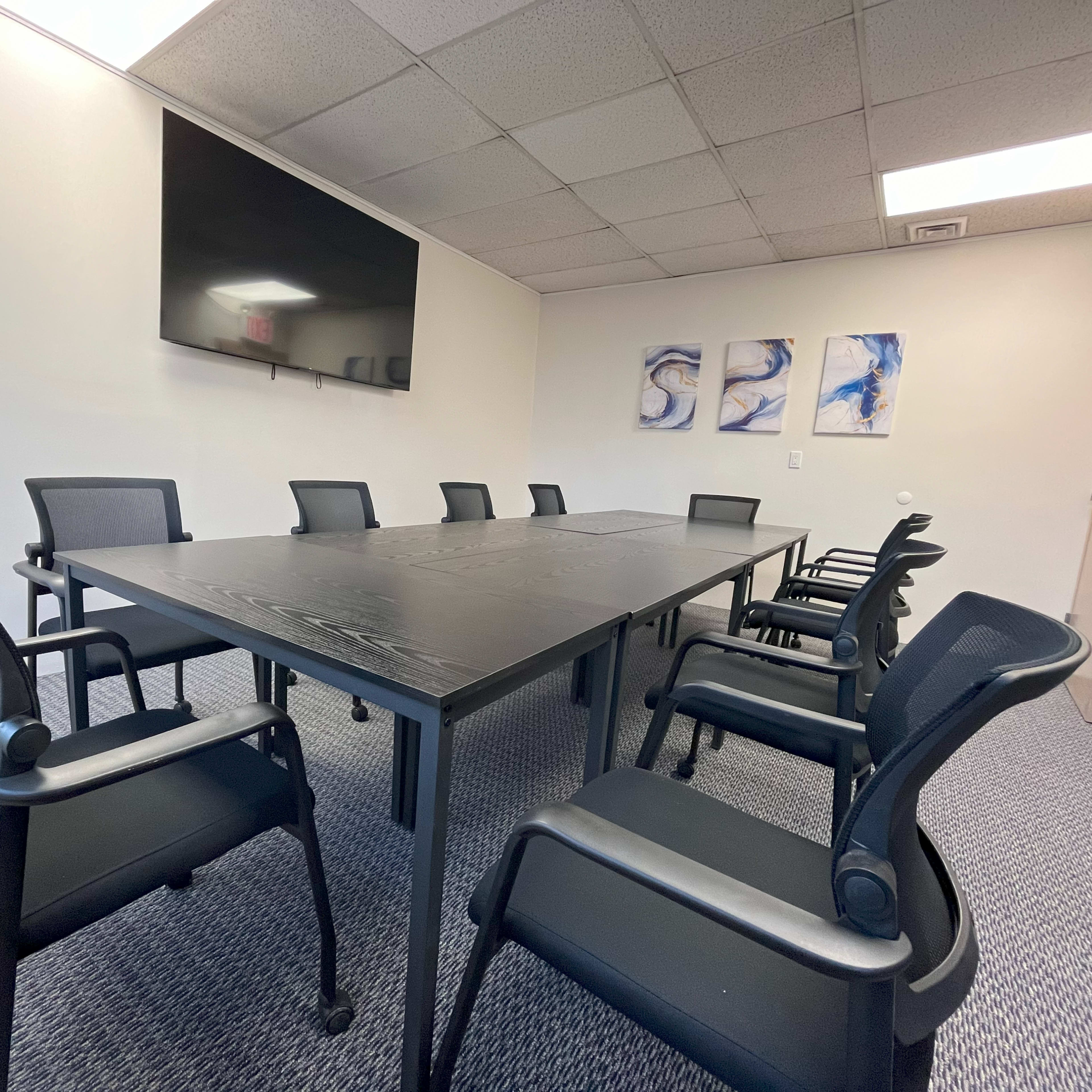 A conference room is set up with a large black table and eight black chairs, featuring a mounted television and three abstract paintings on the wall.