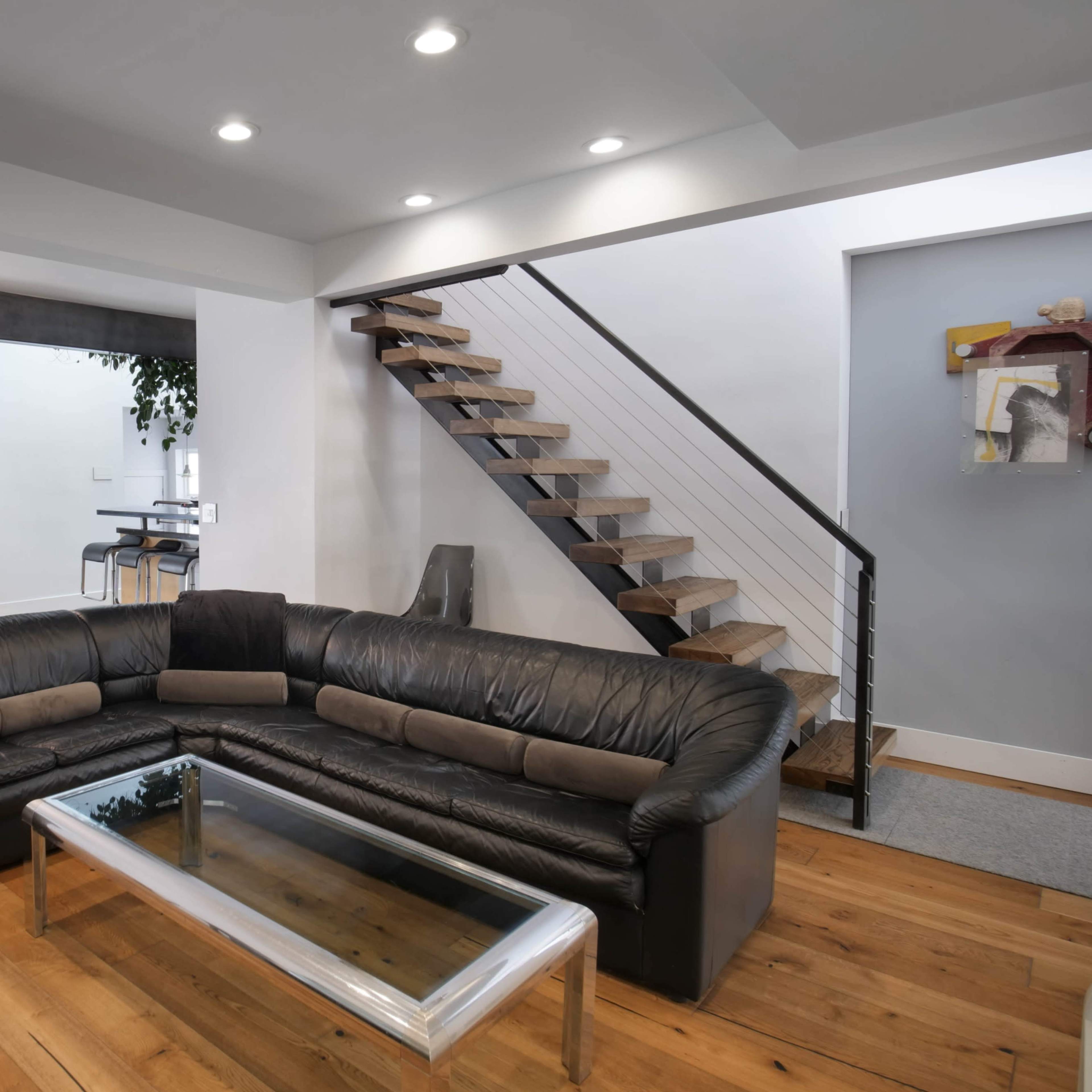 A modern living room with a large black leather sectional sofa, a glass coffee table, and a staircase leading to an upper level.