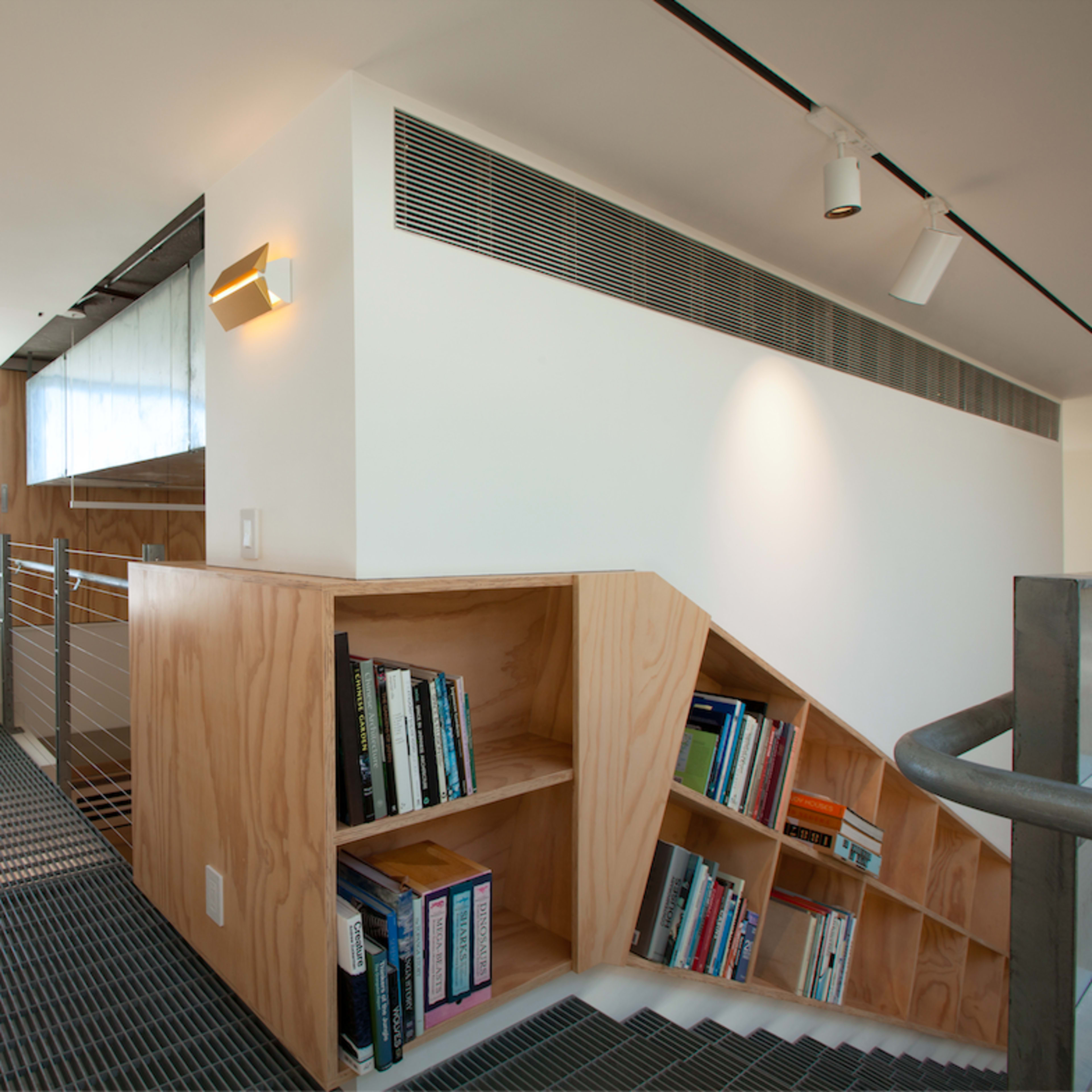 A modern interior space with light-colored walls, a staircase with wooden bookshelves on one side, and a large window that offers a view of the sea.