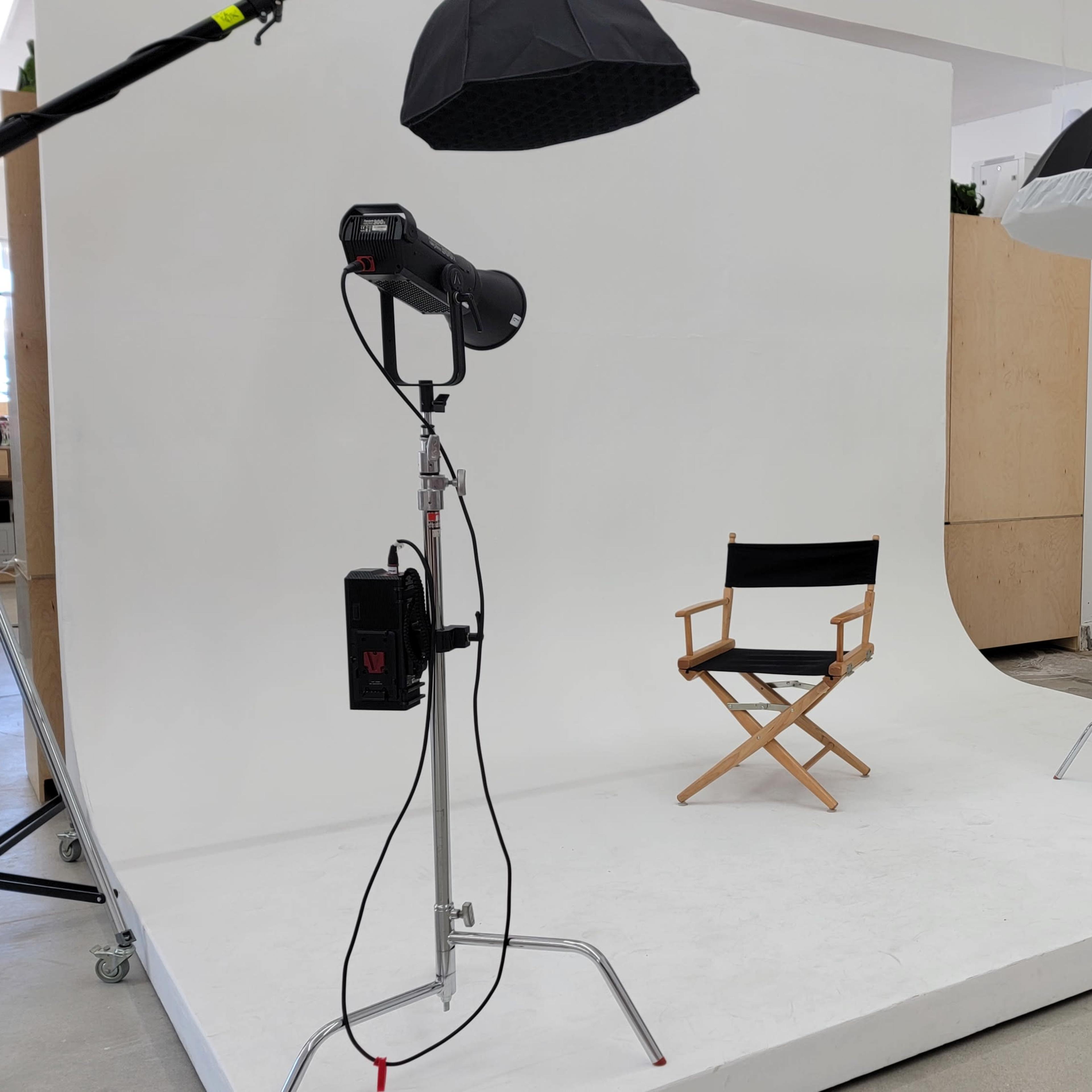 A photography studio setup features a director's chair on a white platform, flanked by two lighting umbrellas and a spotlight.
