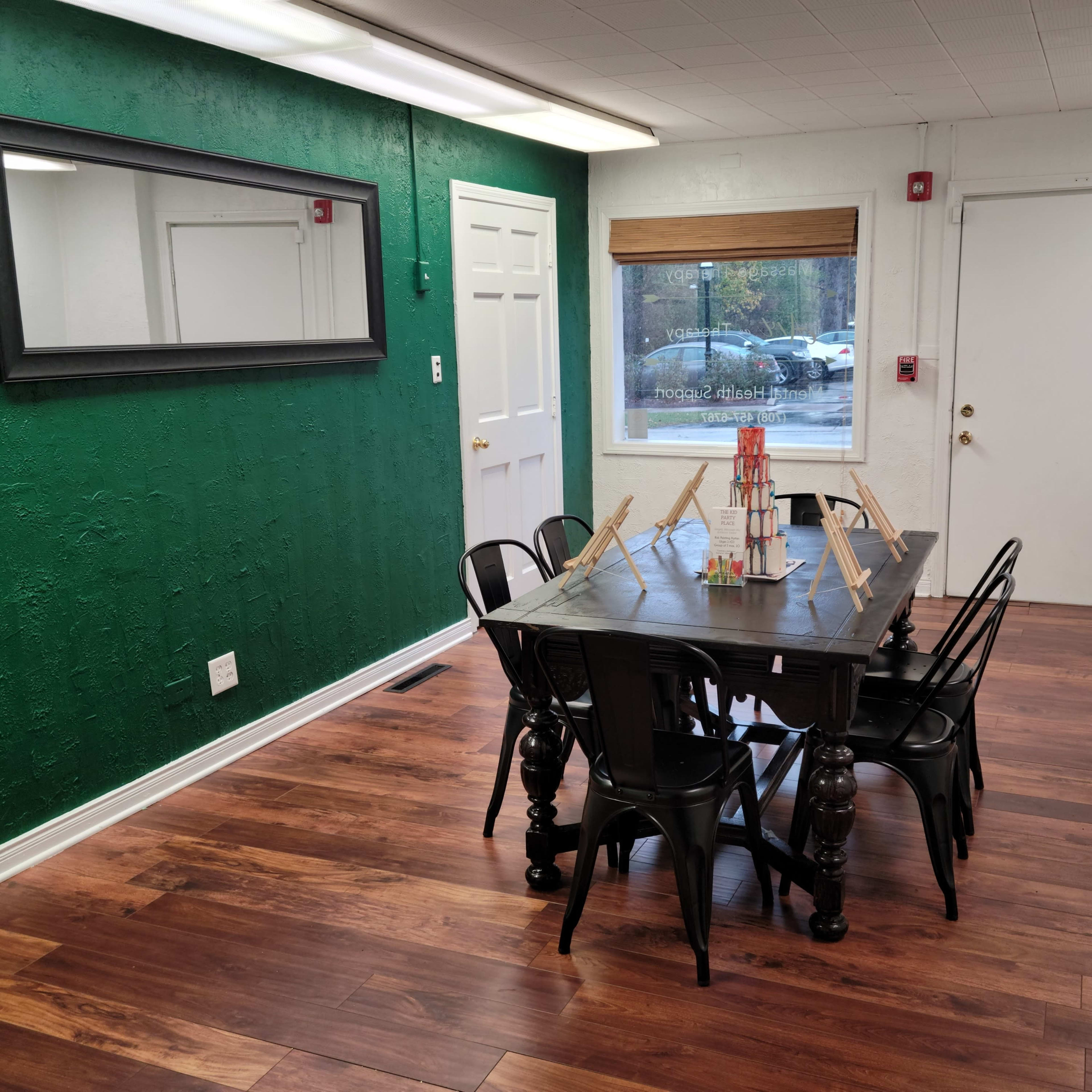 A room with a green accent wall, a large black dining table surrounded by black chairs, and a window with blinds letting in natural light.