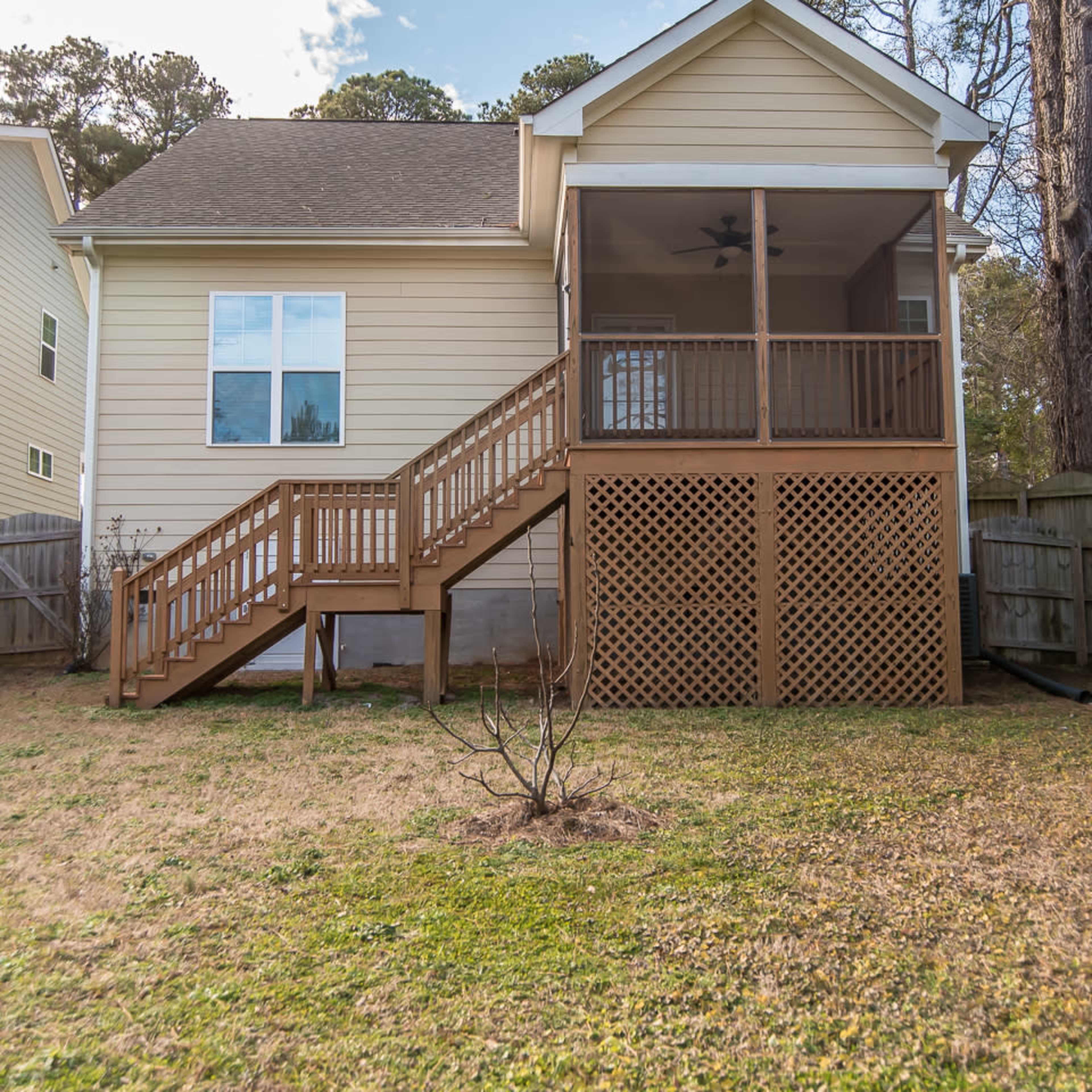 A wooden staircase leads to a screened porch on the back of a two-story house, surrounded by a fenced yard.