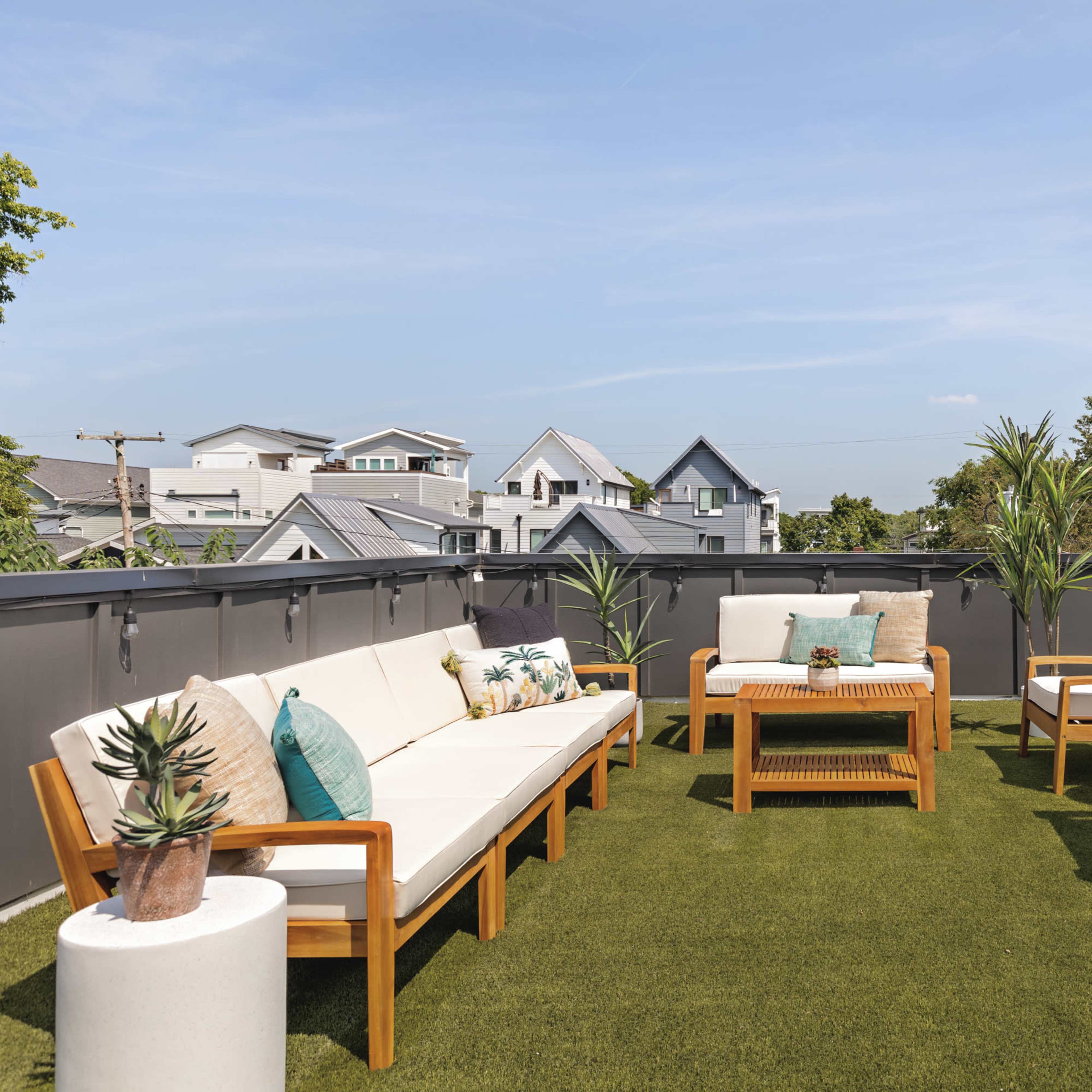 A rooftop patio features outdoor seating with wooden furniture, potted plants, and a carpet of artificial grass, overlooking residential buildings and green trees.