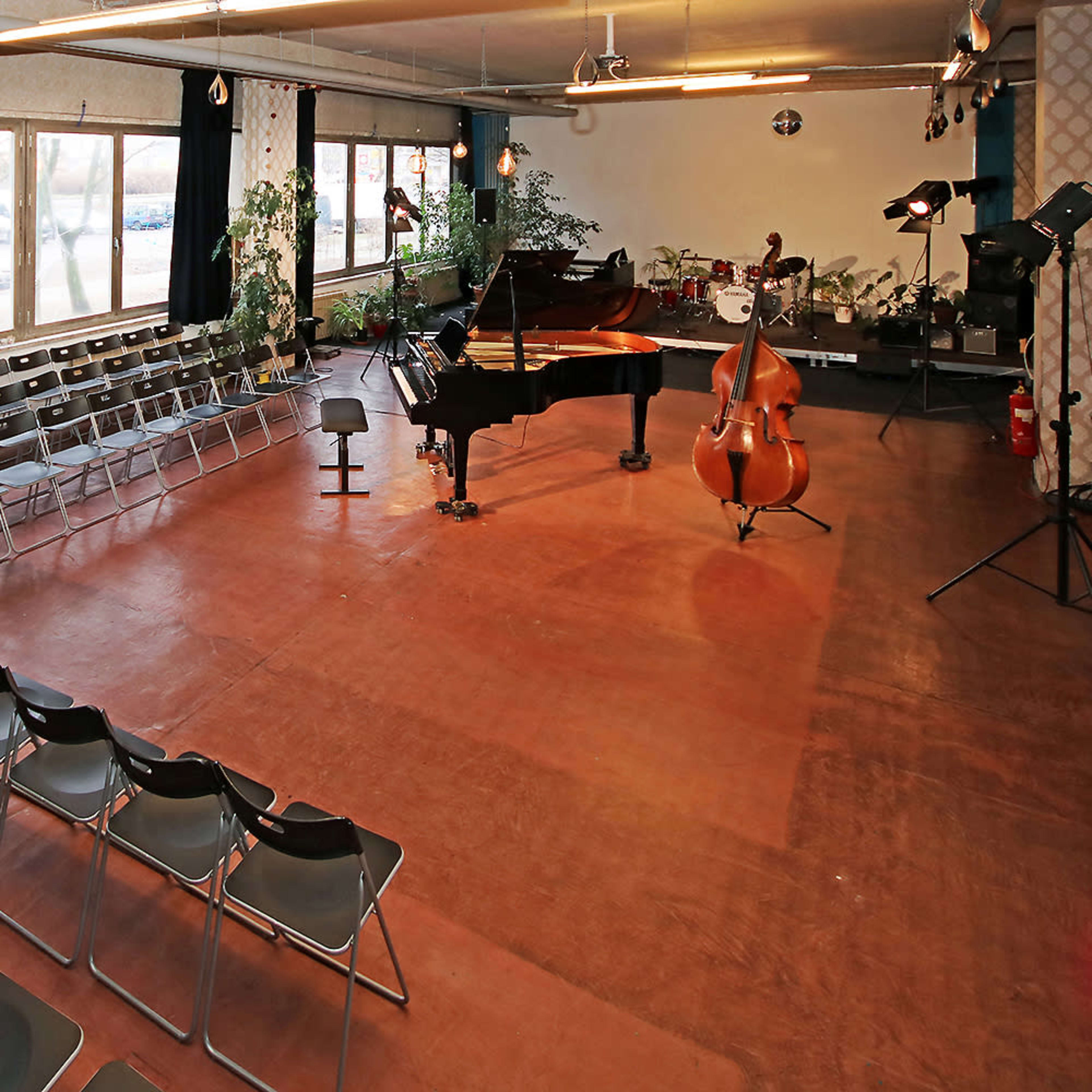 A music performance space features rows of black chairs facing a grand piano and double bass, with large windows providing natural light.