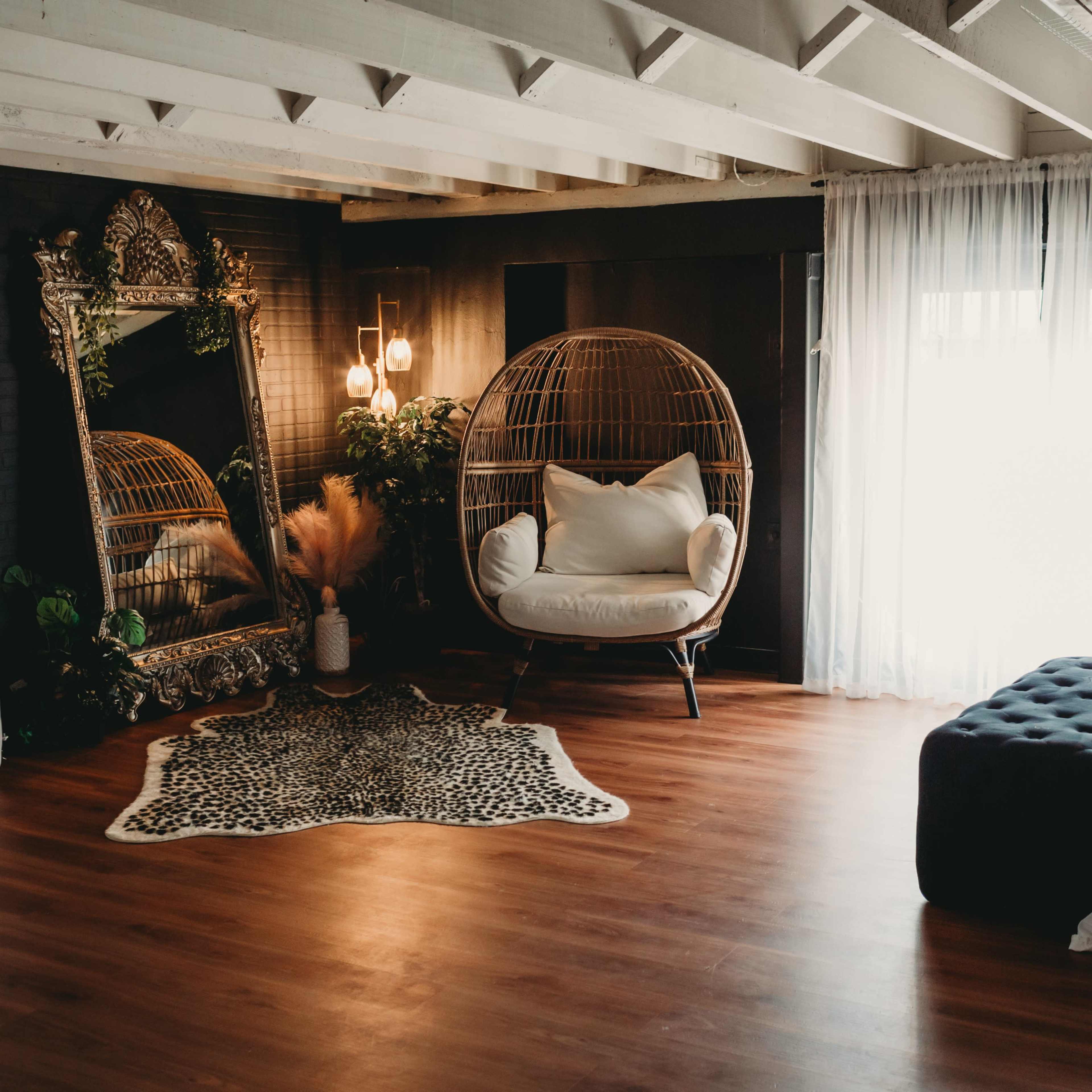 A spacious indoor area with wooden flooring, featuring a large decorative mirror, a hanging chair, and a plush ottoman, all surrounded by sheer white curtains and a decorative rug.