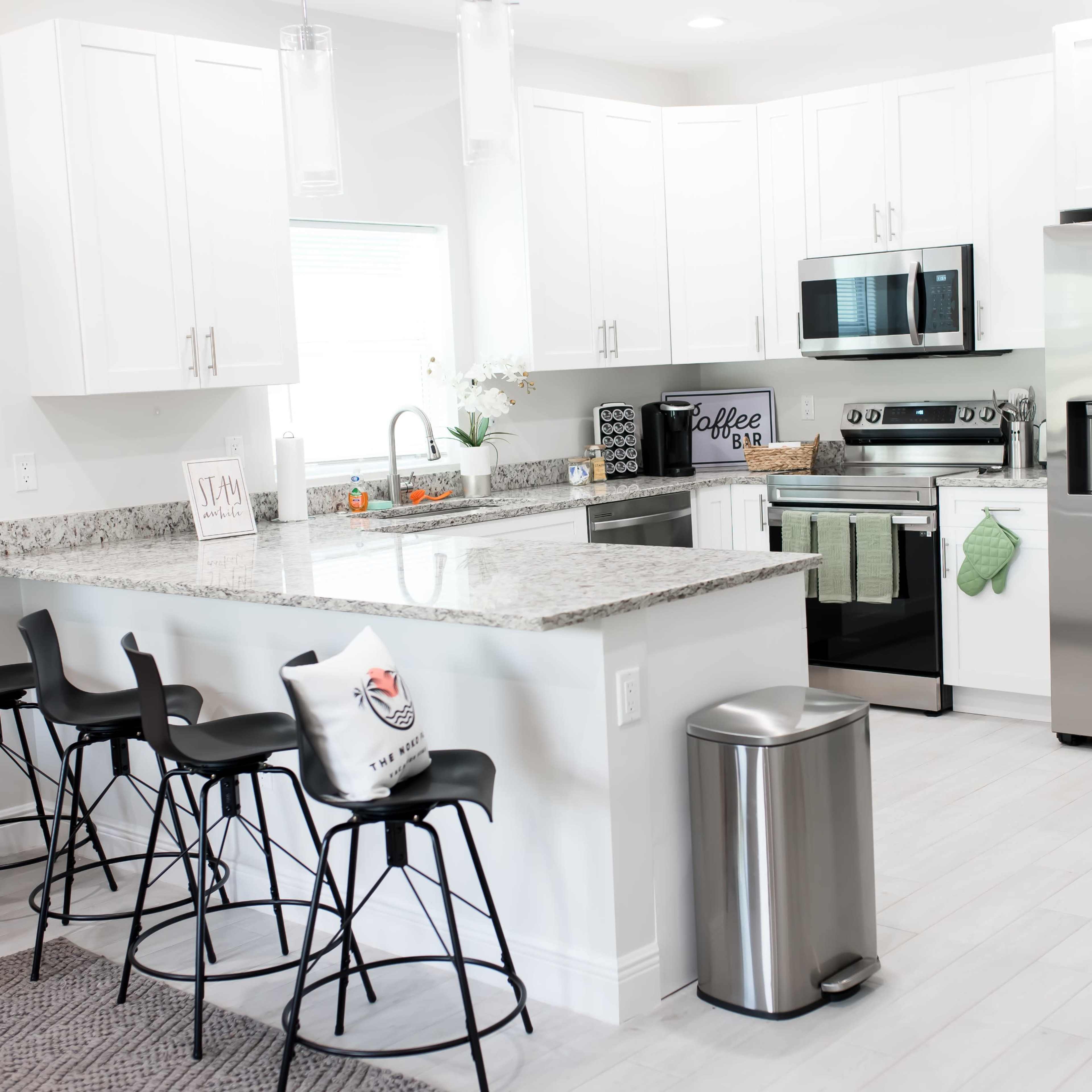 A modern kitchen features white cabinets, stainless steel appliances, and a granite countertop with bar stools at a breakfast bar.