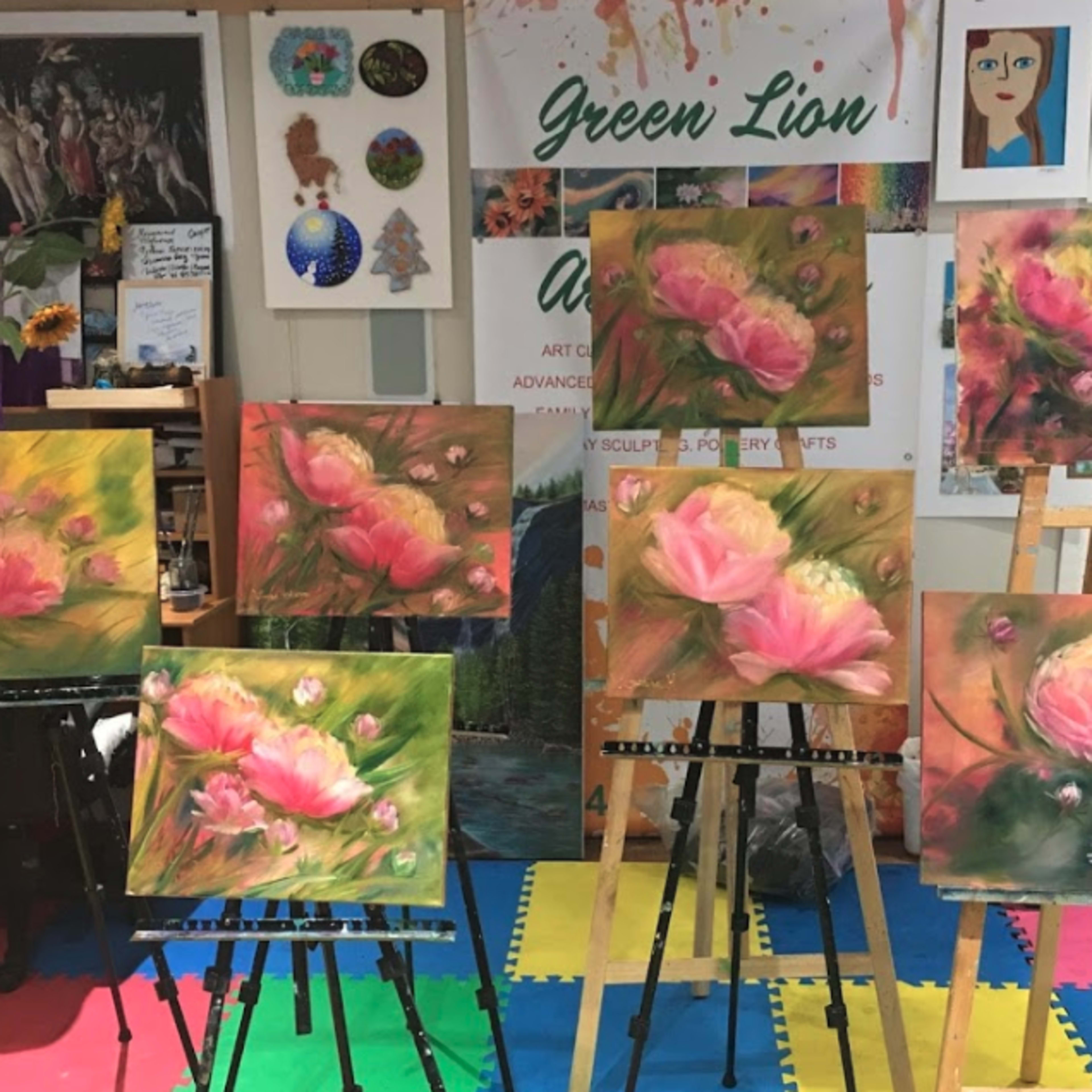 Several flower paintings displayed on easels in an art studio with colorful flooring and various artworks on the walls.