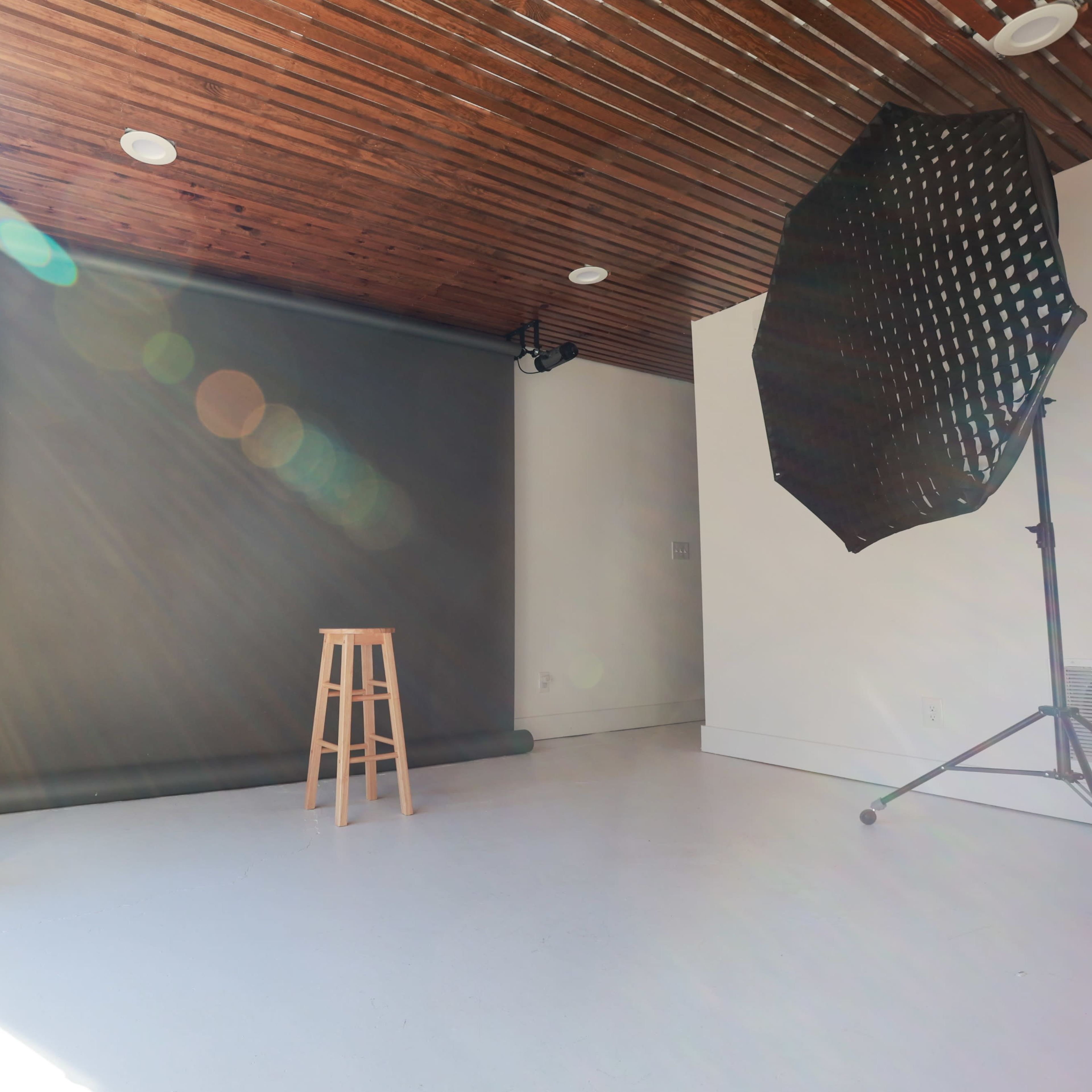 A minimalist photography studio features a wooden stool, a potted plant, and a large softbox near a gray backdrop.