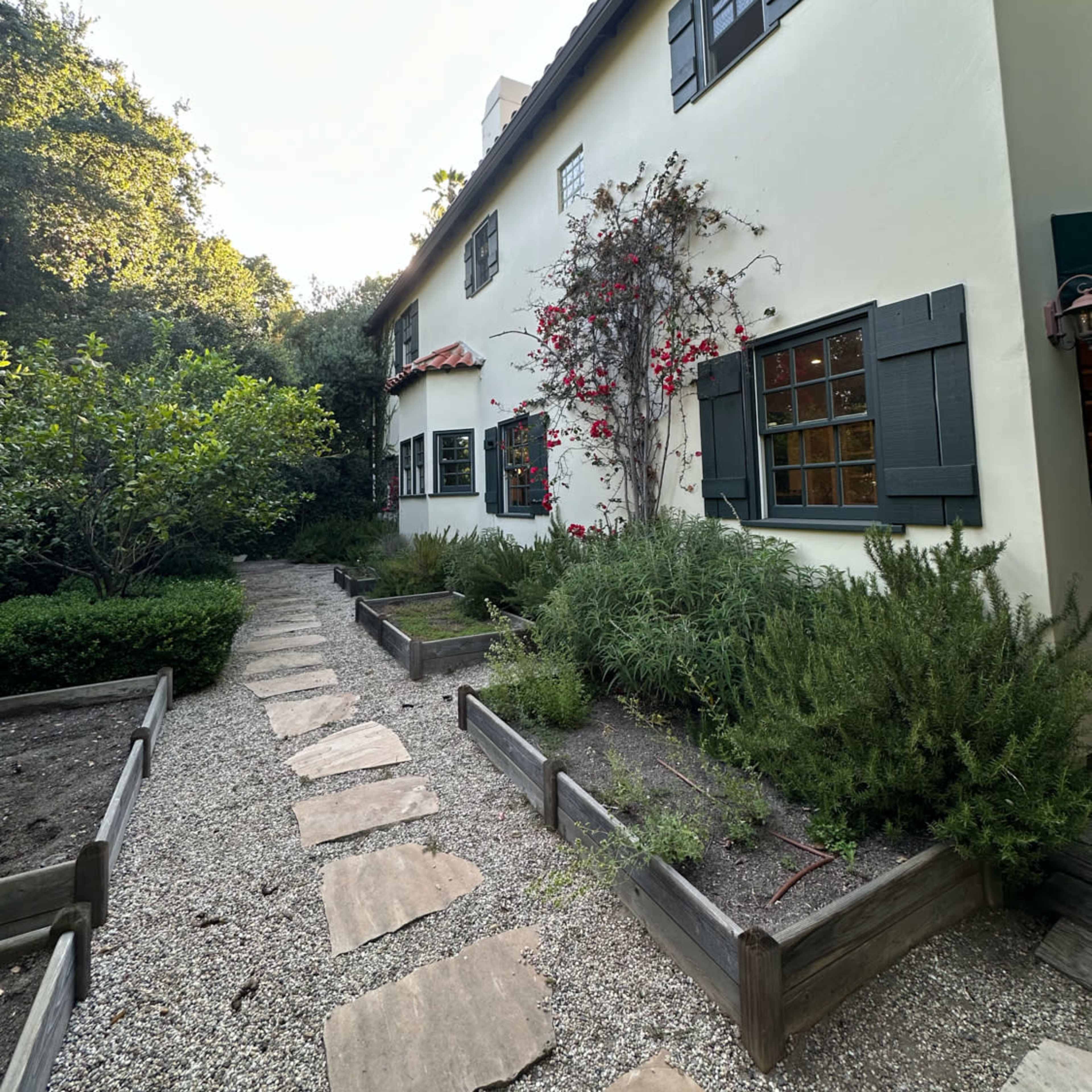 A stone pathway leads past landscaped garden beds to a two-story white house with dark green shutters and a climbing plant on its exterior.
