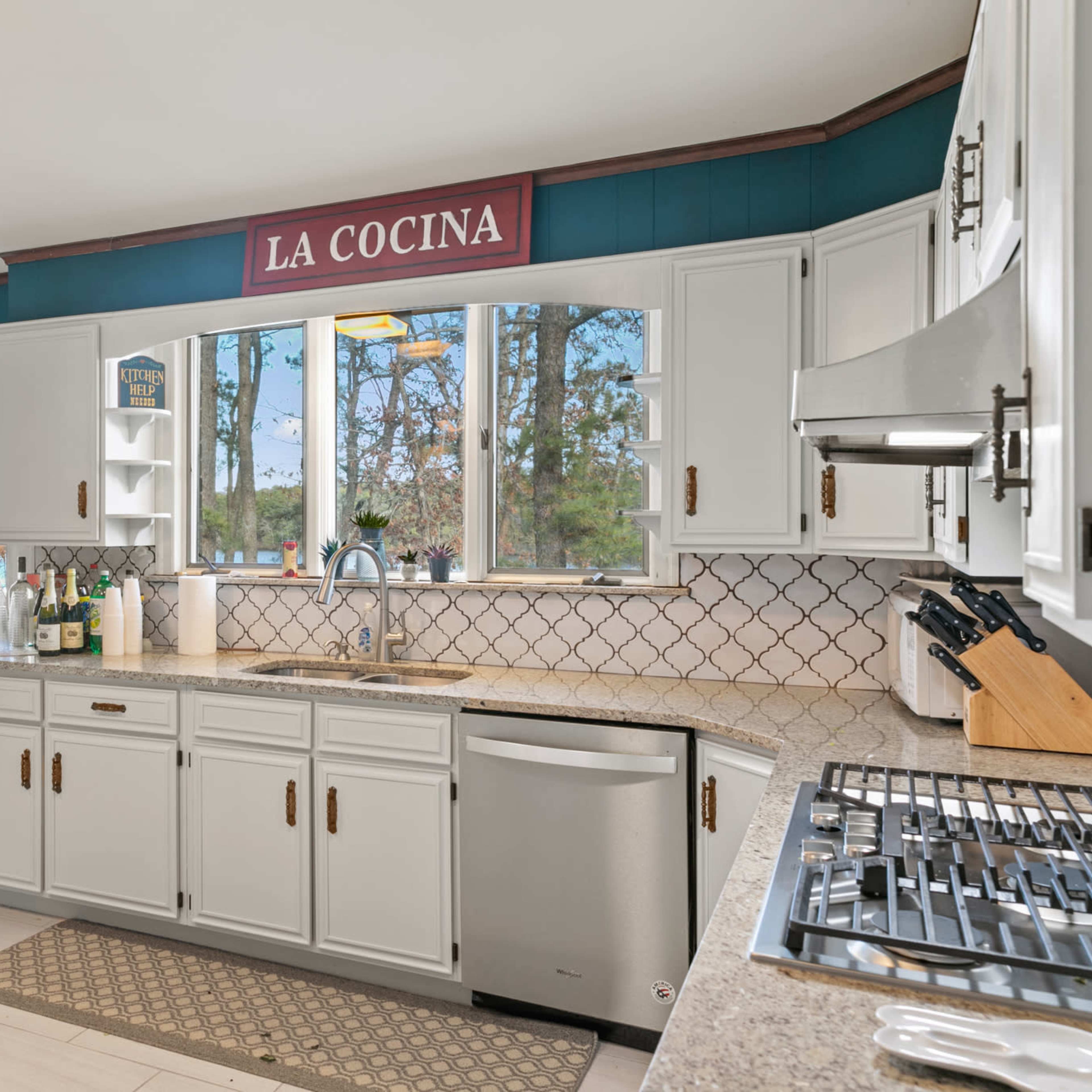 A bright kitchen features white cabinets, a tiled backsplash, and large windows overlooking a natural landscape.