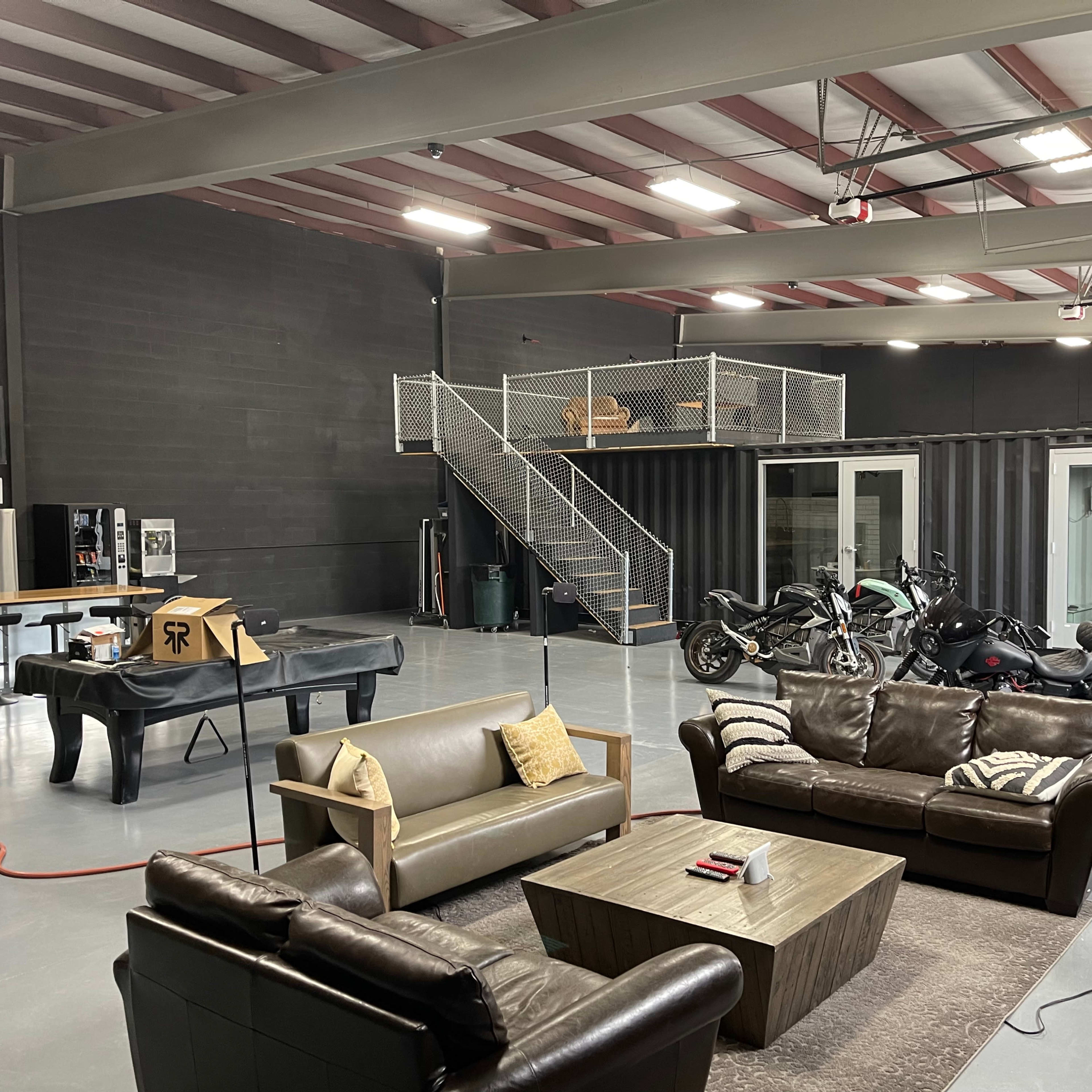 A spacious, industrial-style interior featuring a seating area with leather sofas, a coffee table, and a piano, along with a two-wheeled motorcycle parked nearby and a staircase leading to a mezzanine level.