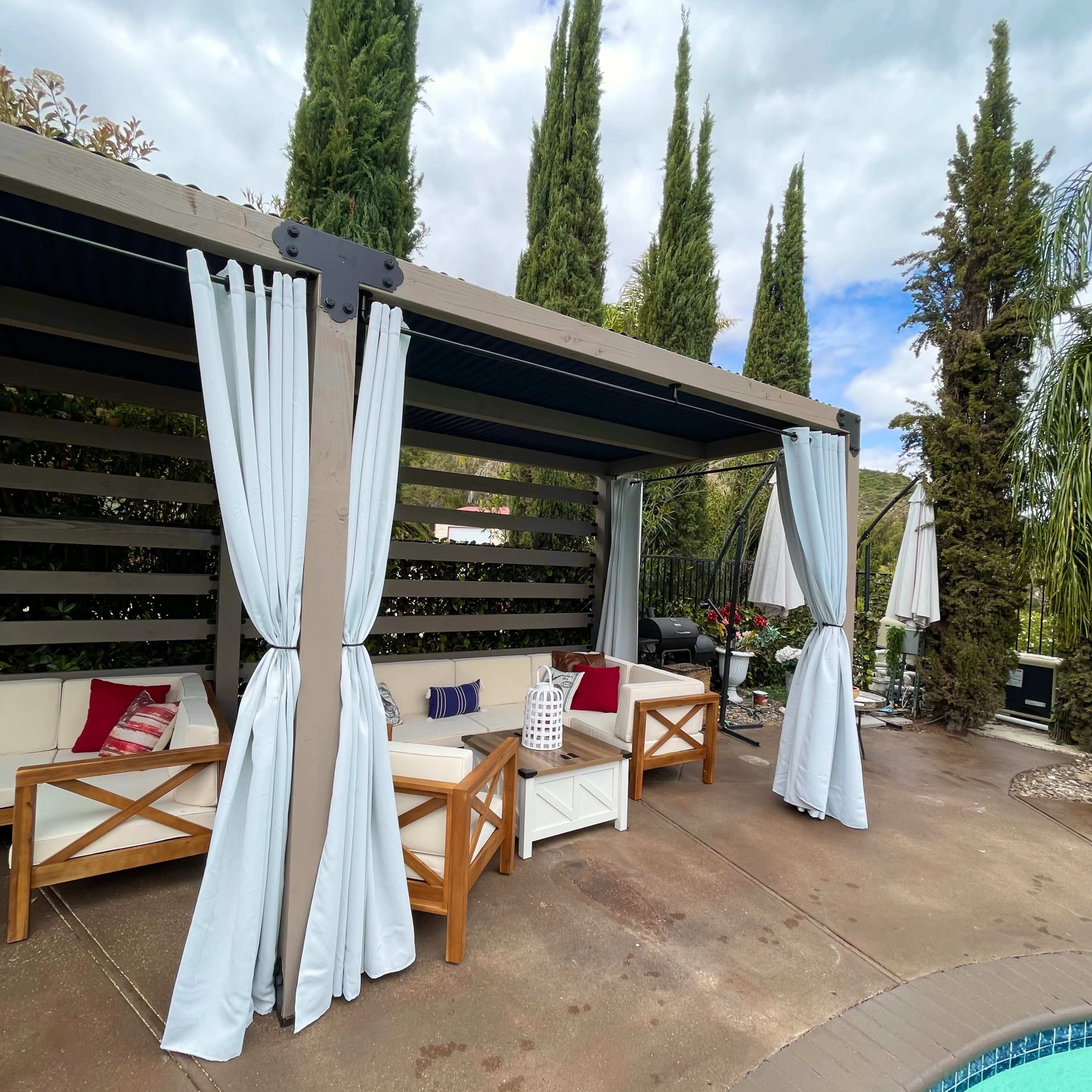 A covered outdoor lounge area with curtains and seating is situated beside a swimming pool, surrounded by tall greenery.