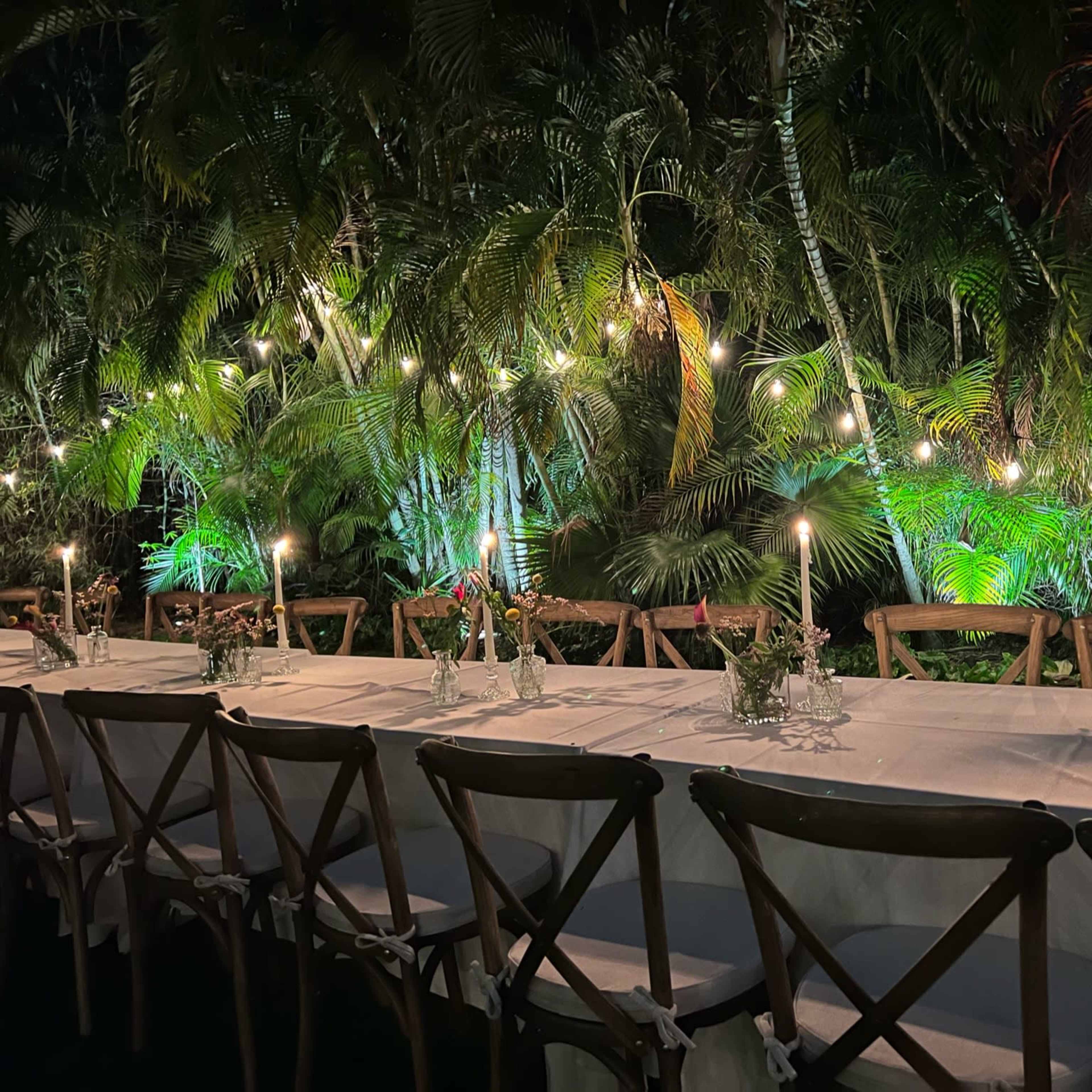 A long dining table is set outdoors, surrounded by tropical plants and illuminated by soft candlelight and string lights.