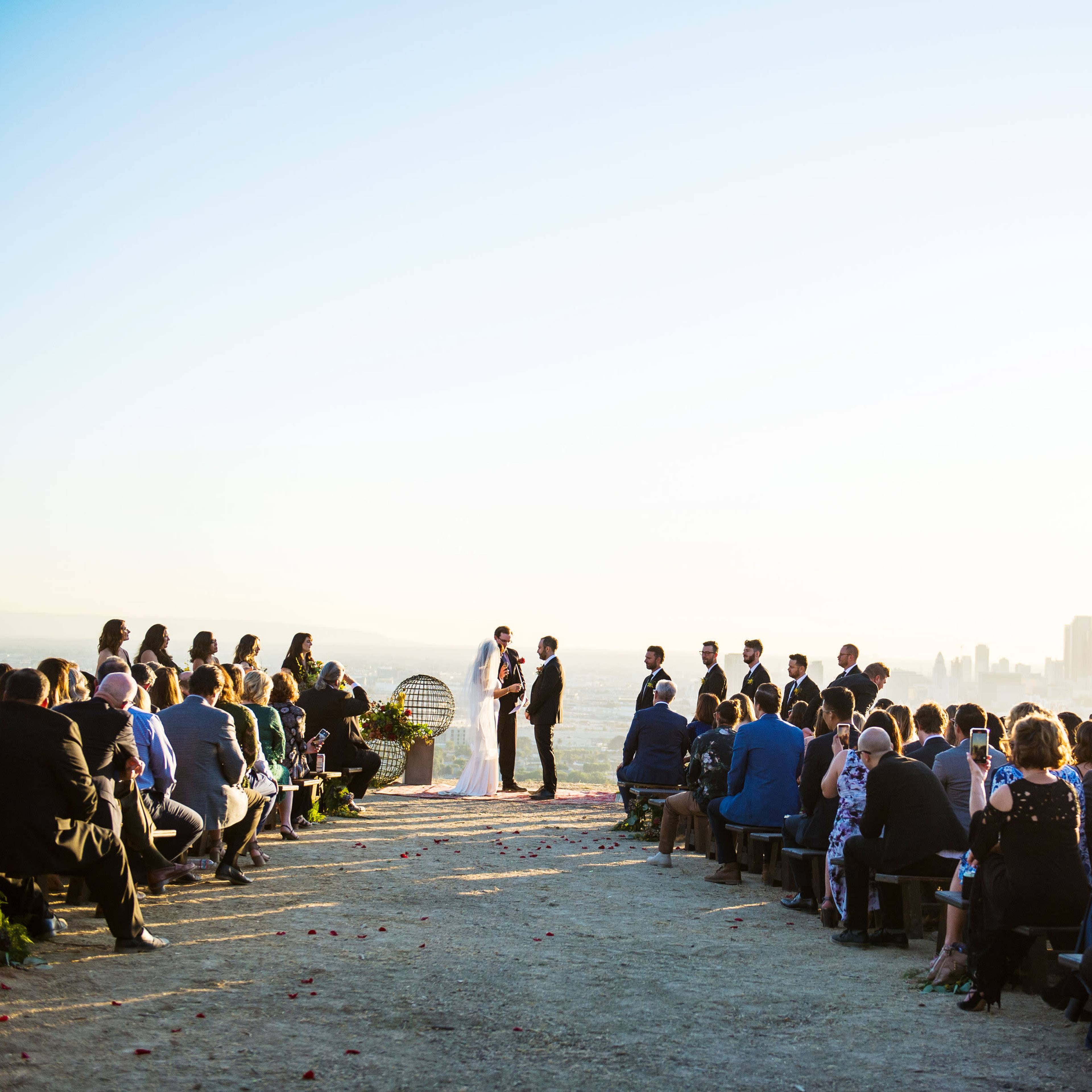 A couple exchanges vows during an outdoor wedding ceremony with a city skyline in the background and guests seated on wooden benches.