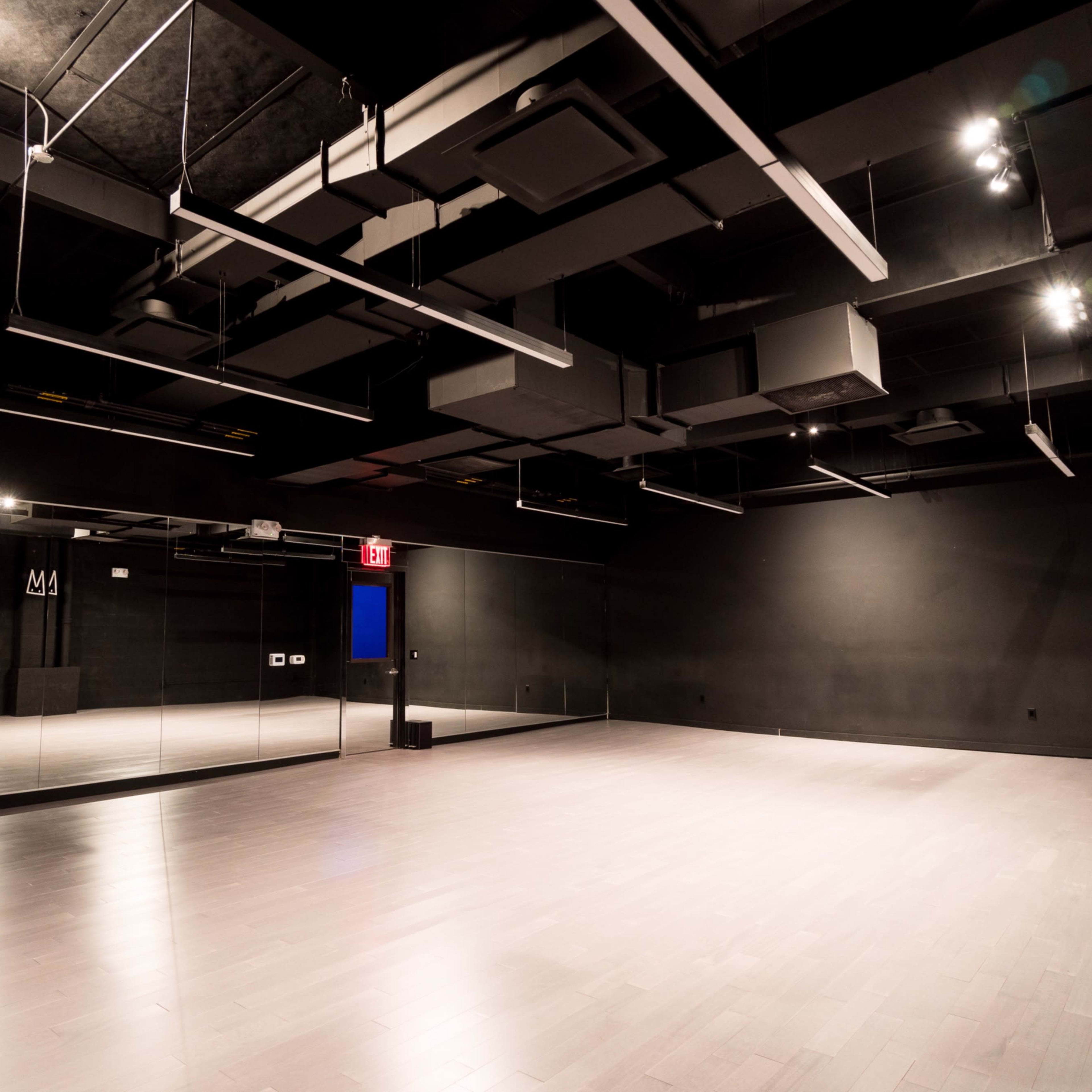 An empty dance studio featuring wooden flooring, black walls, and a large mirror on one side, with overhead lighting.