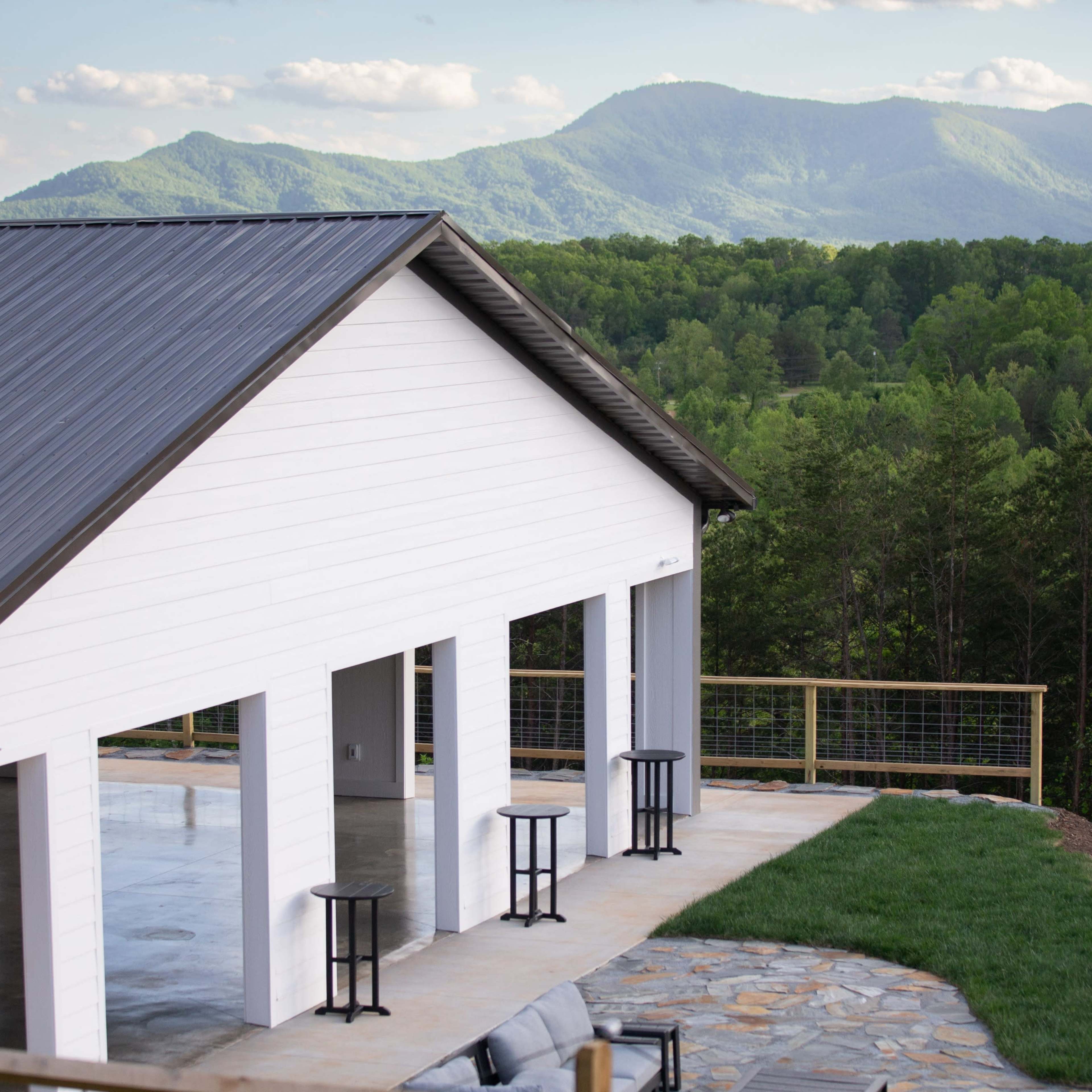 A modern white building with a metal roof overlooks a mountainous landscape, featuring expansive greenery and a stone patio with black stools.