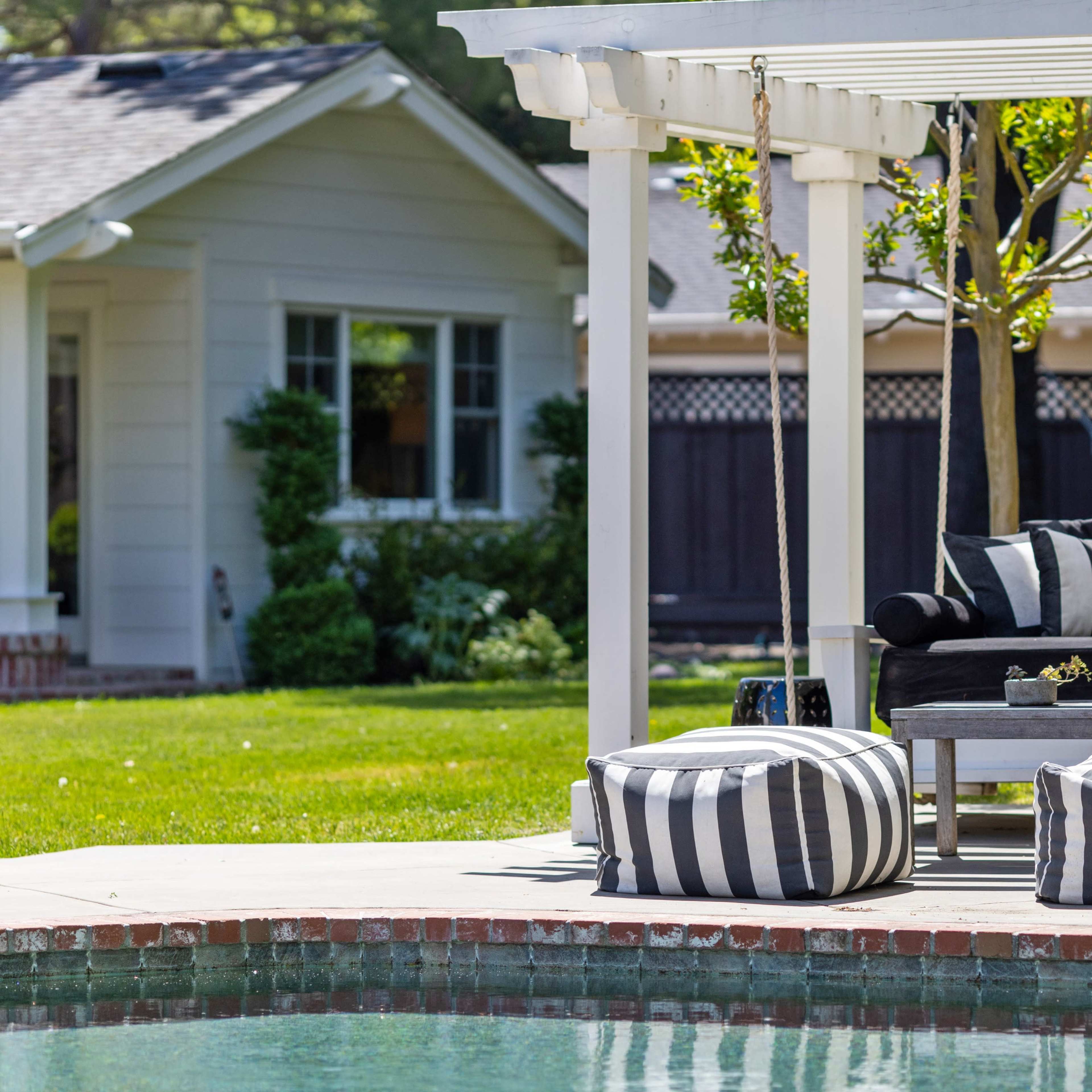A covered patio with black and white striped cushions overlooks a swimming pool and a grassy yard, with a house in the background.
