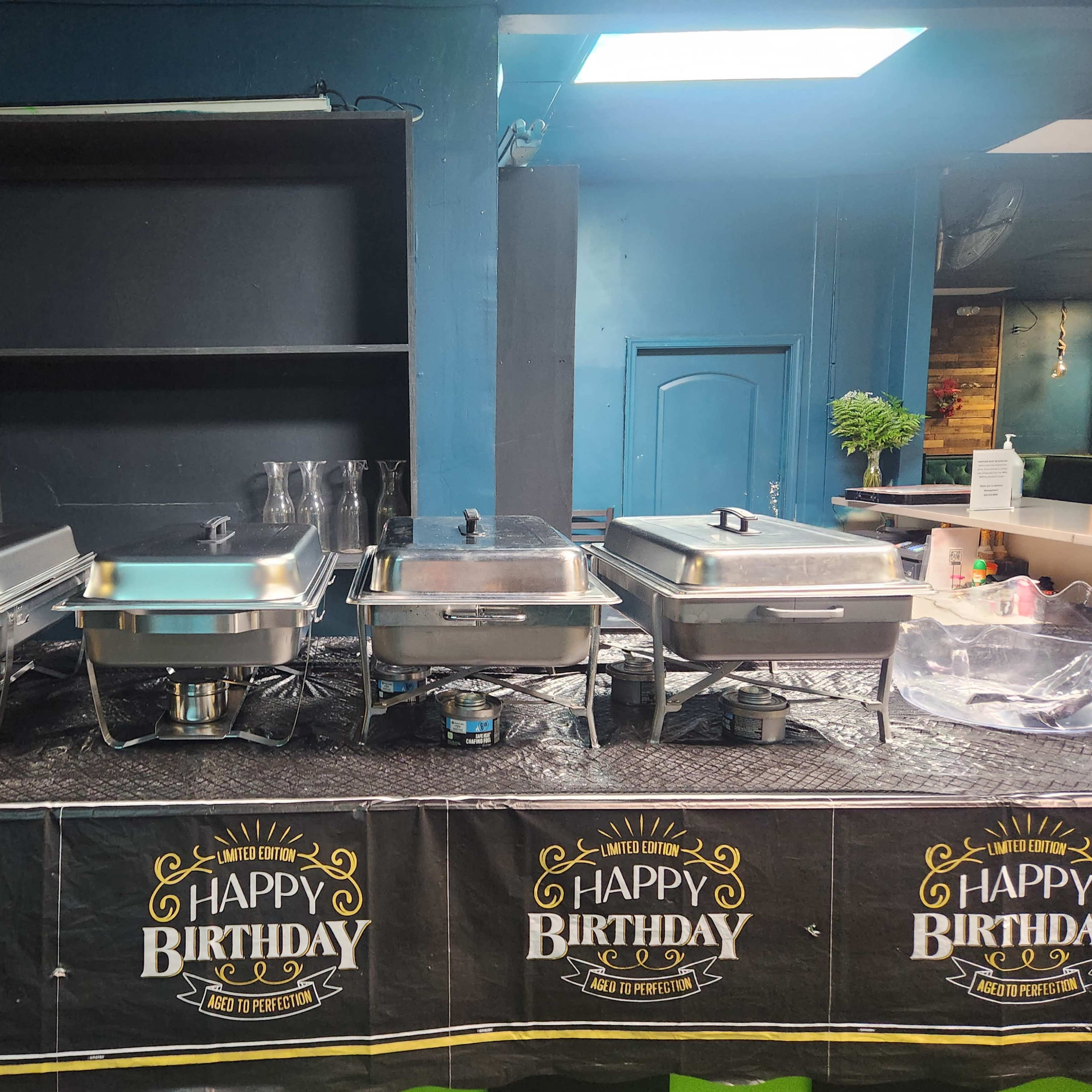 A row of covered chafing dishes is displayed on a table adorned with a birthday banner.