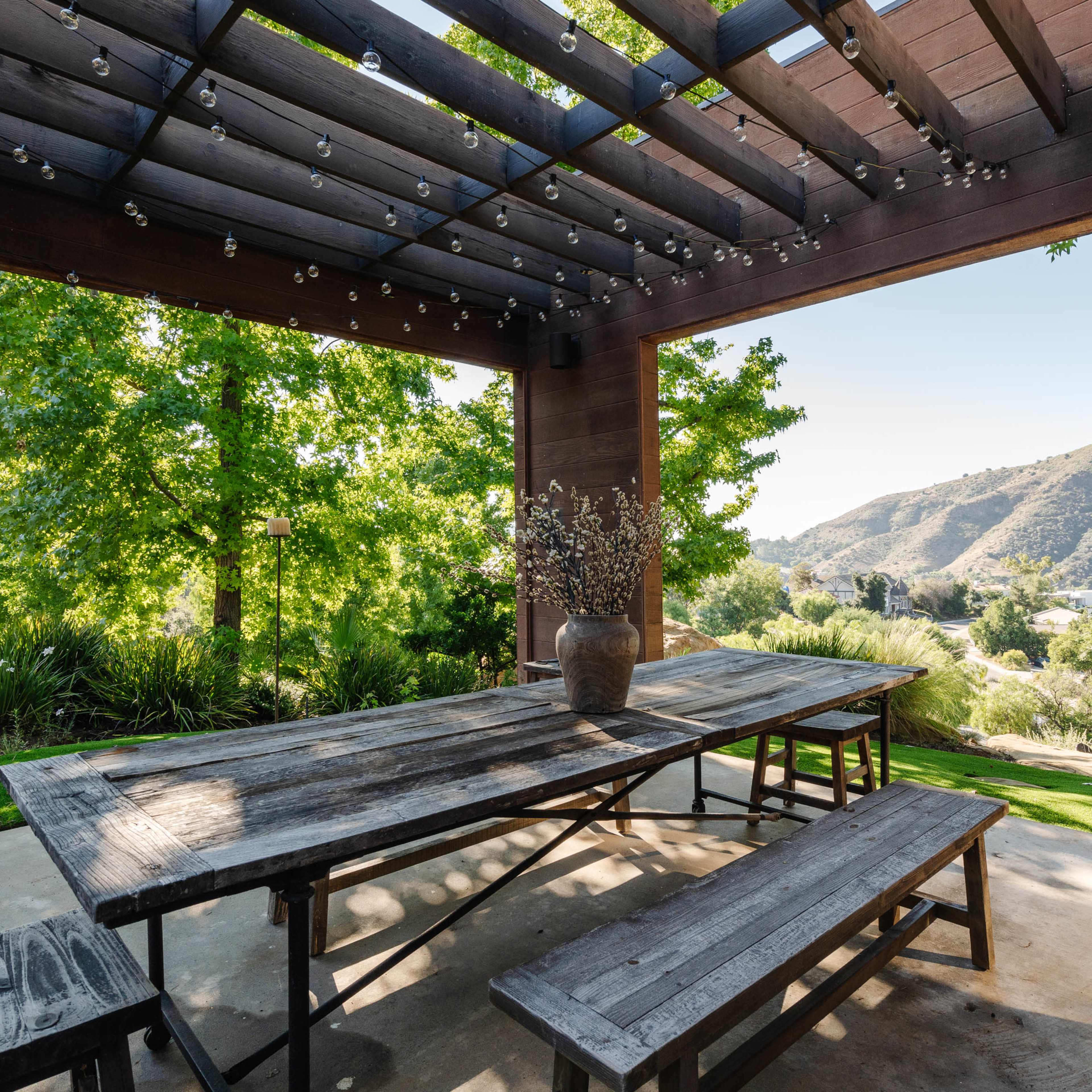 A wooden picnic table with benches sits under a pergola, surrounded by greenery and offering a view of rolling hills in the background.