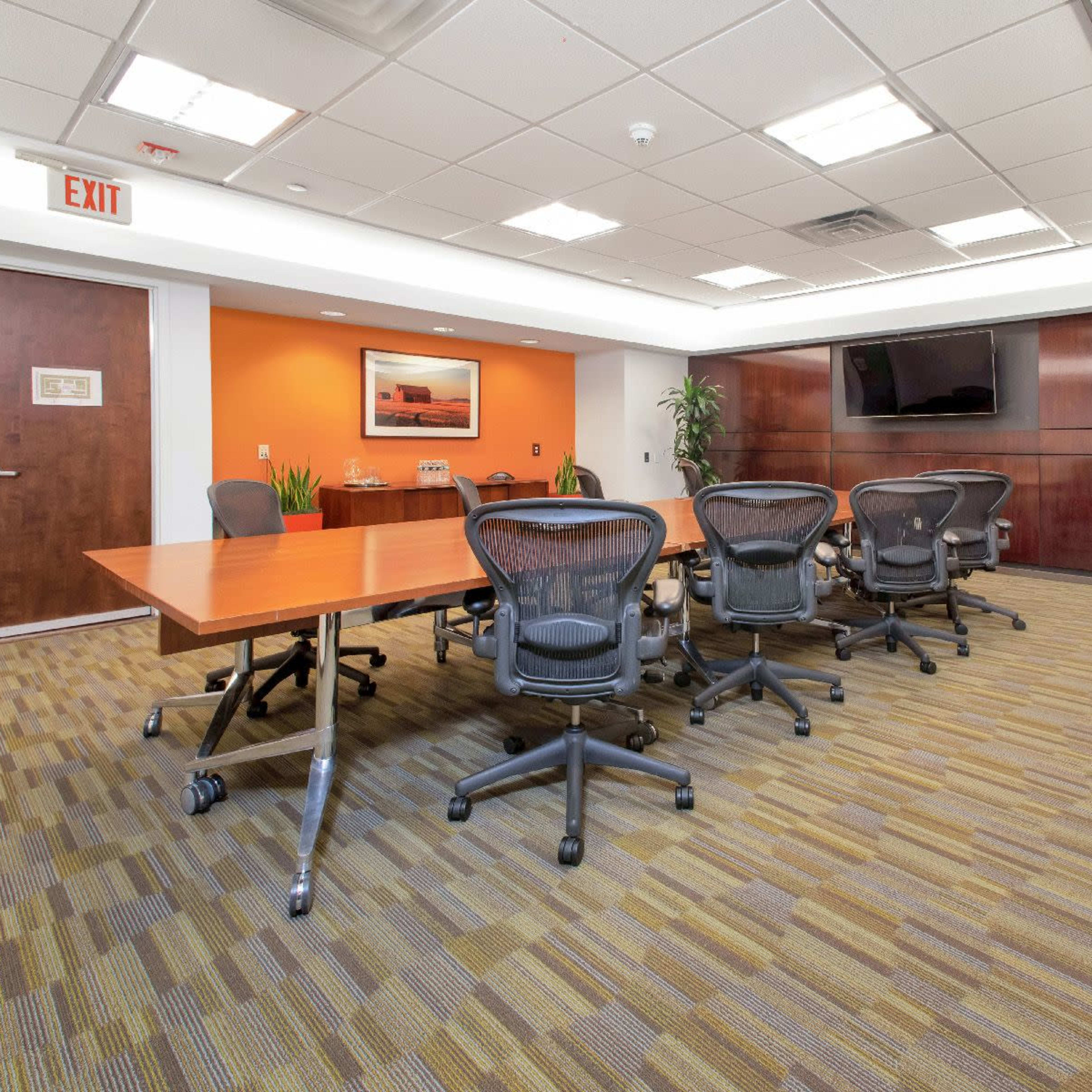 A conference room features a long wooden table surrounded by rolling office chairs, with an orange accent wall and a large television mounted on the wall.