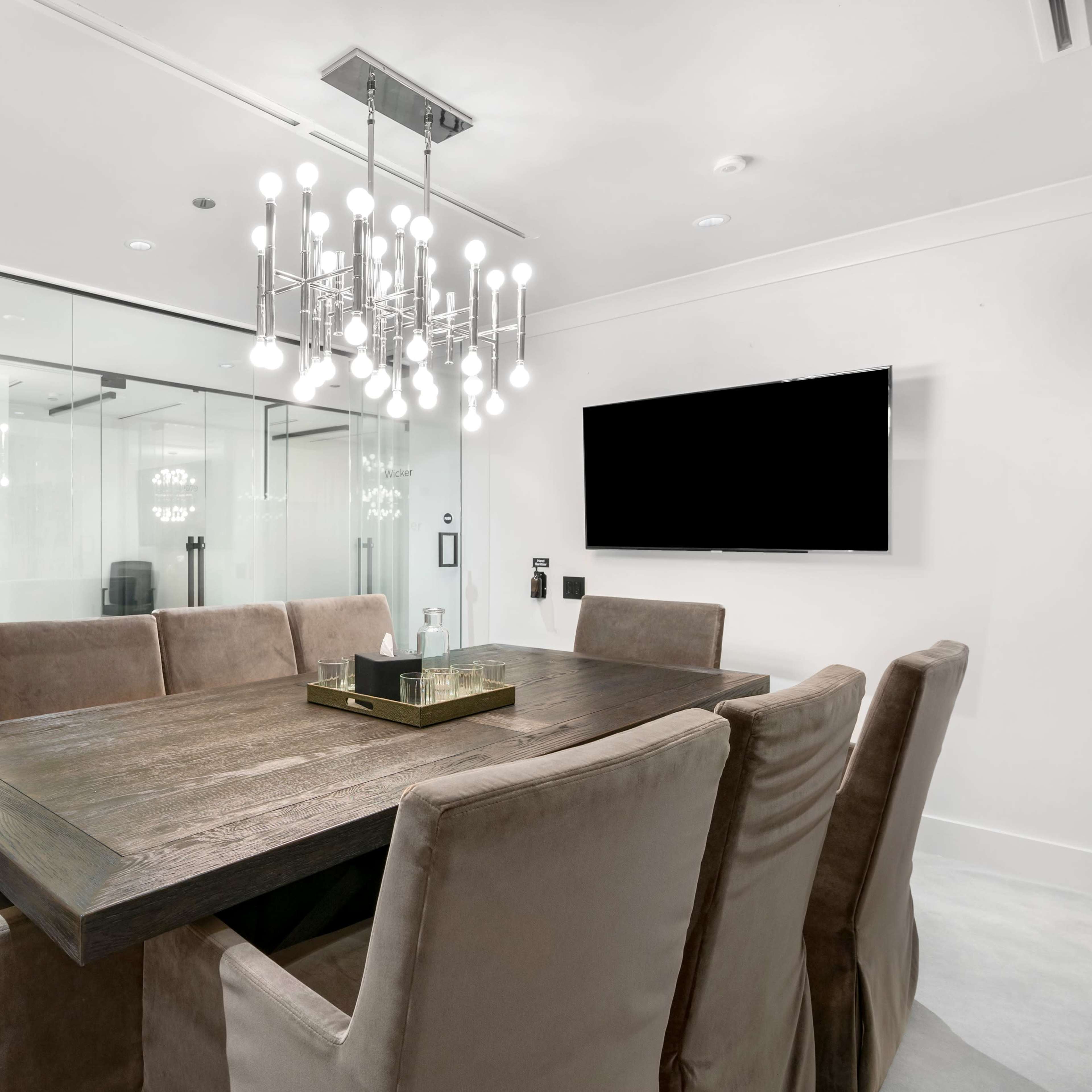 A modern conference room features a wooden table surrounded by eight upholstered chairs, with a glass wall showcasing an adjacent meeting space.