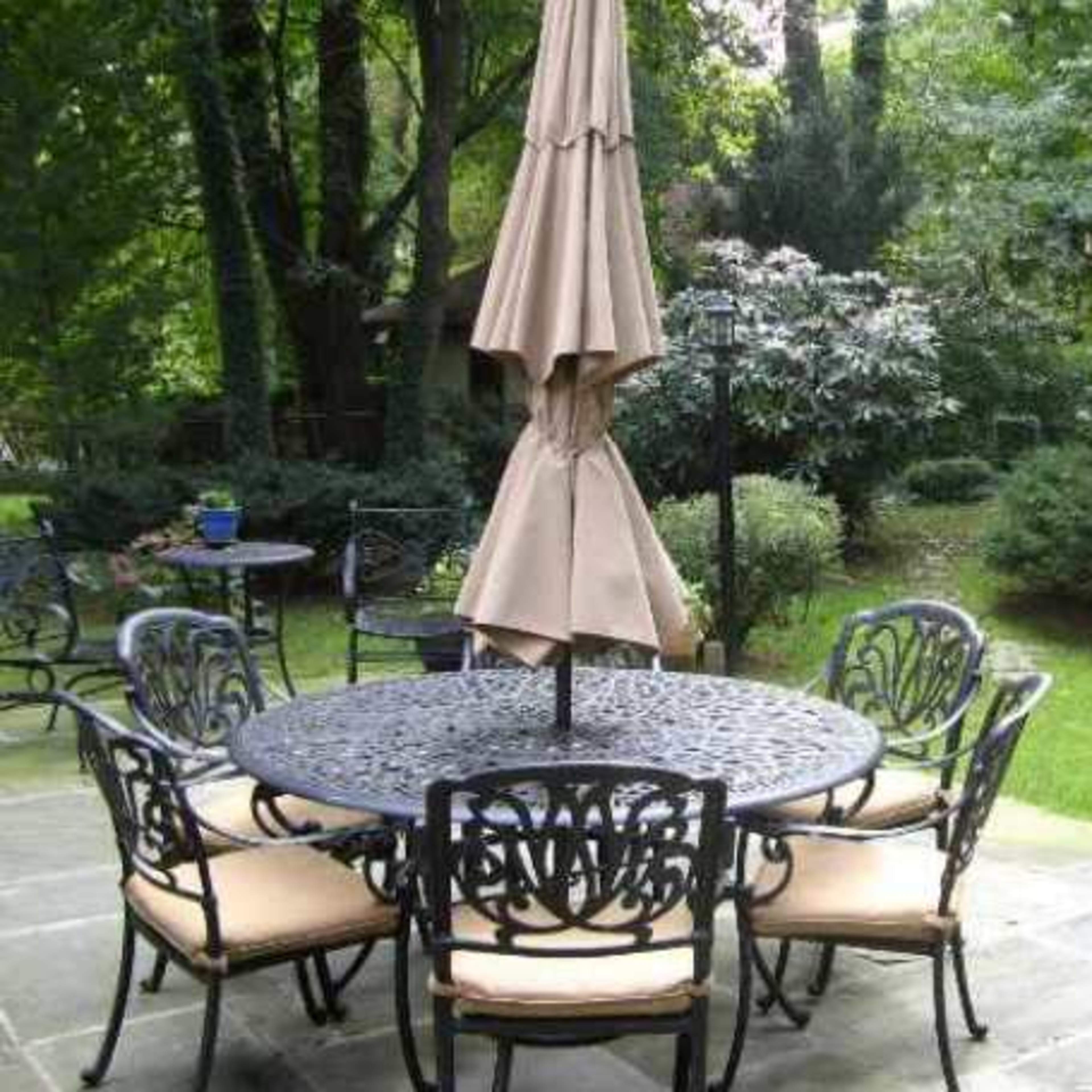 A round metal table with six cushioned chairs is set on a stone patio under a partially opened umbrella, surrounded by greenery.