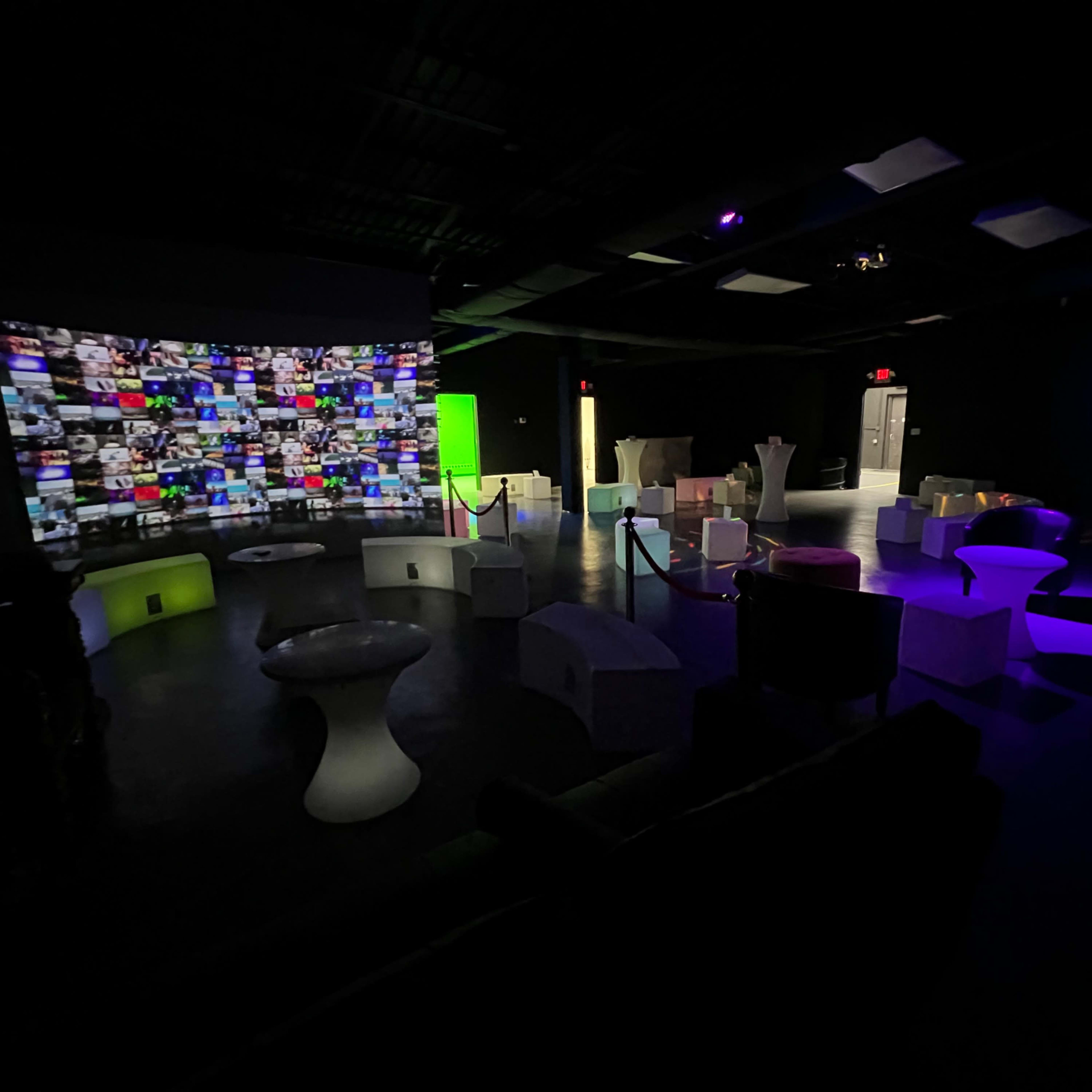 A dimly lit room with various illuminated seating arrangements and a large screen displaying a collage of colorful images on one wall.
