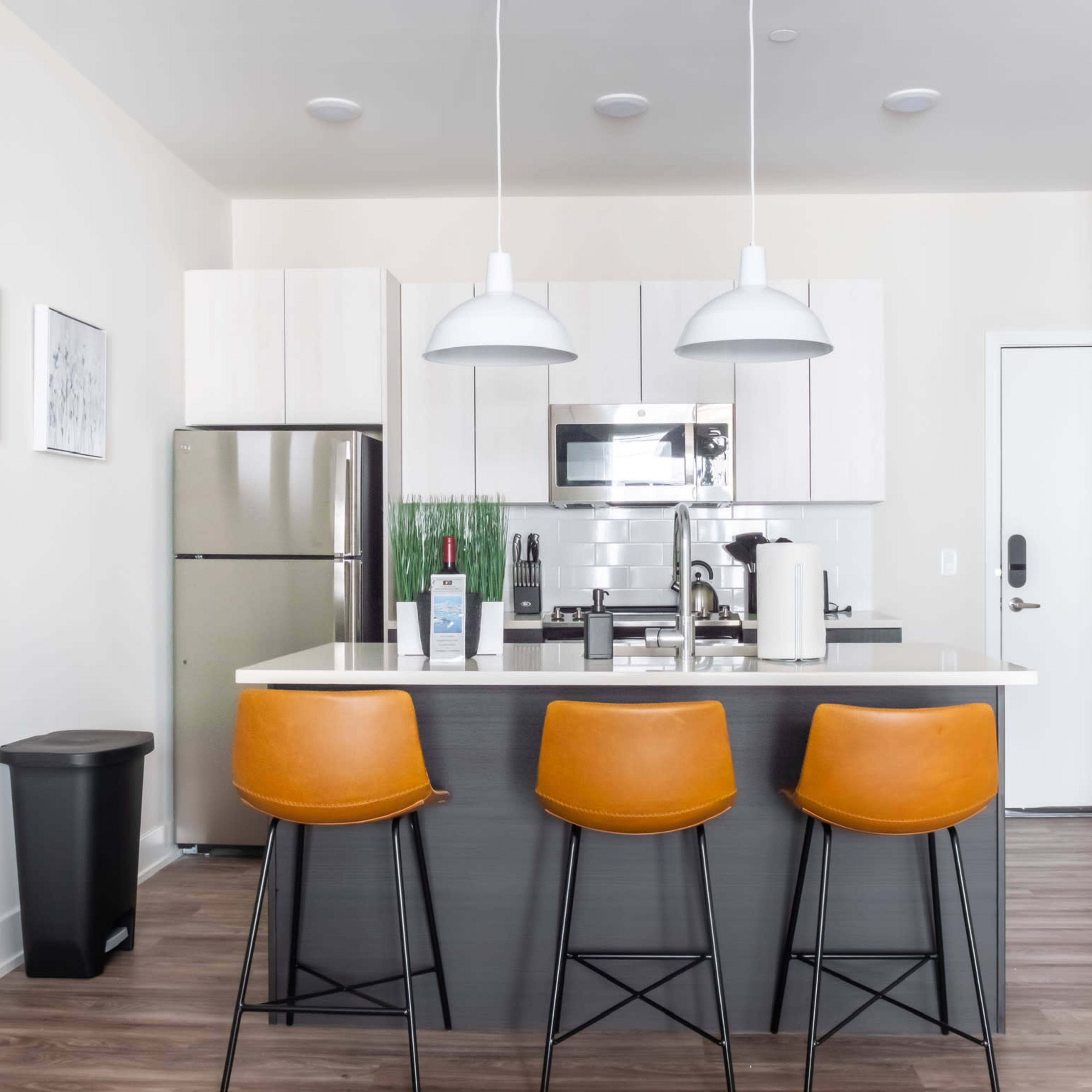 A modern kitchen features a central island with three brown bar stools, stainless steel appliances, and a door leading to a hallway.