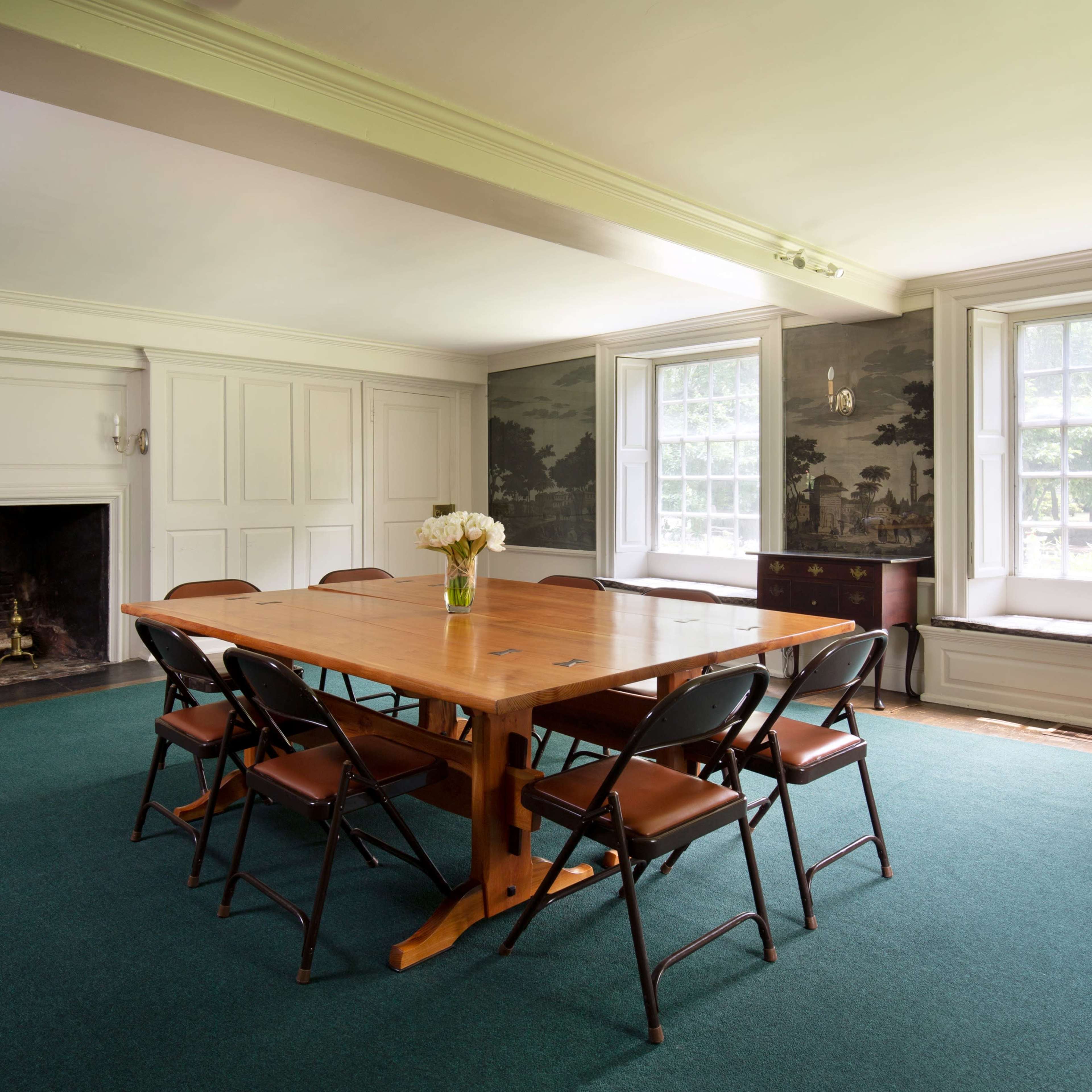 A wooden table surrounded by black chairs is positioned in a room with a fireplace and large windows featuring a mural on the walls.