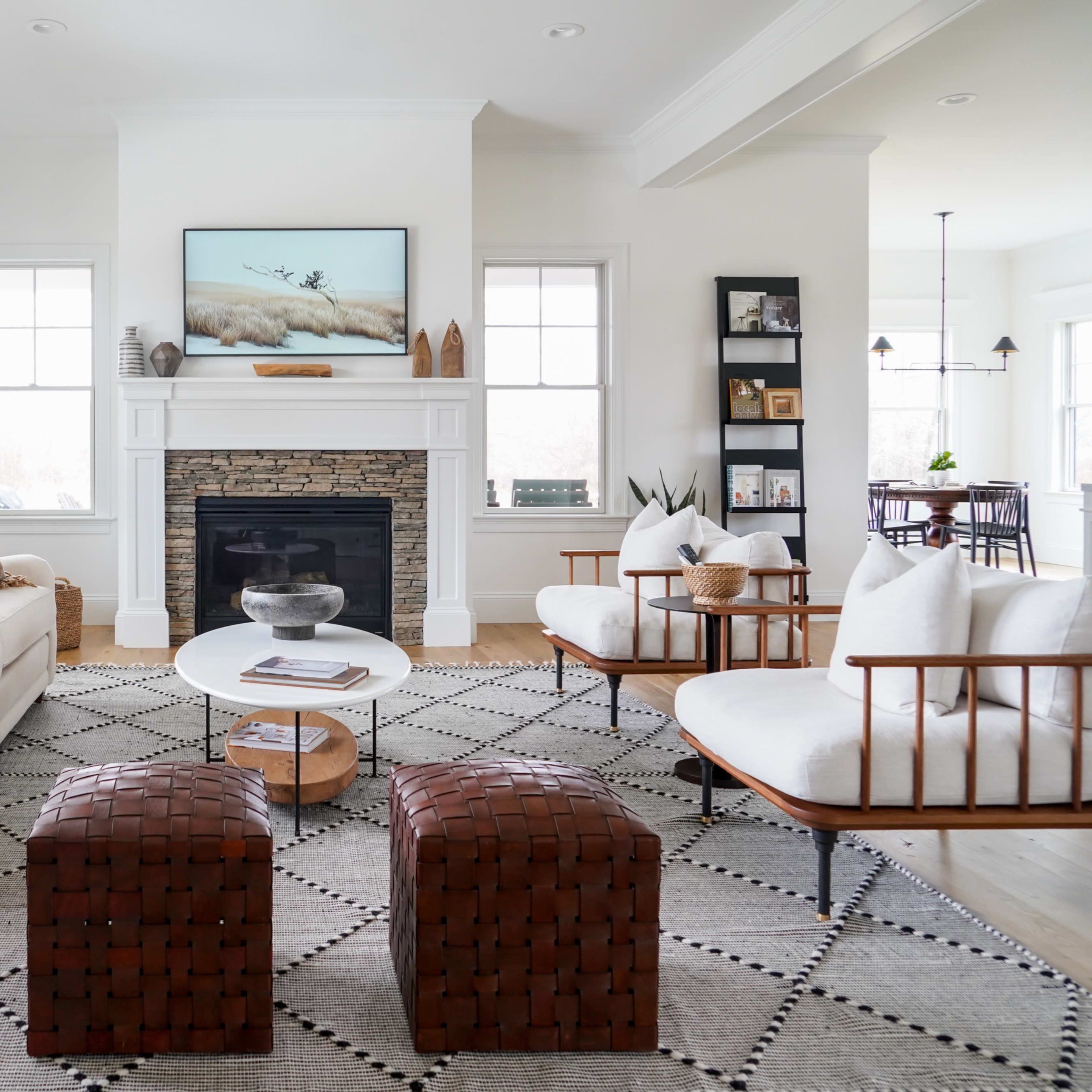 A bright living room with a white sofa, two wooden chairs, a round coffee table, and a fireplace under a television.