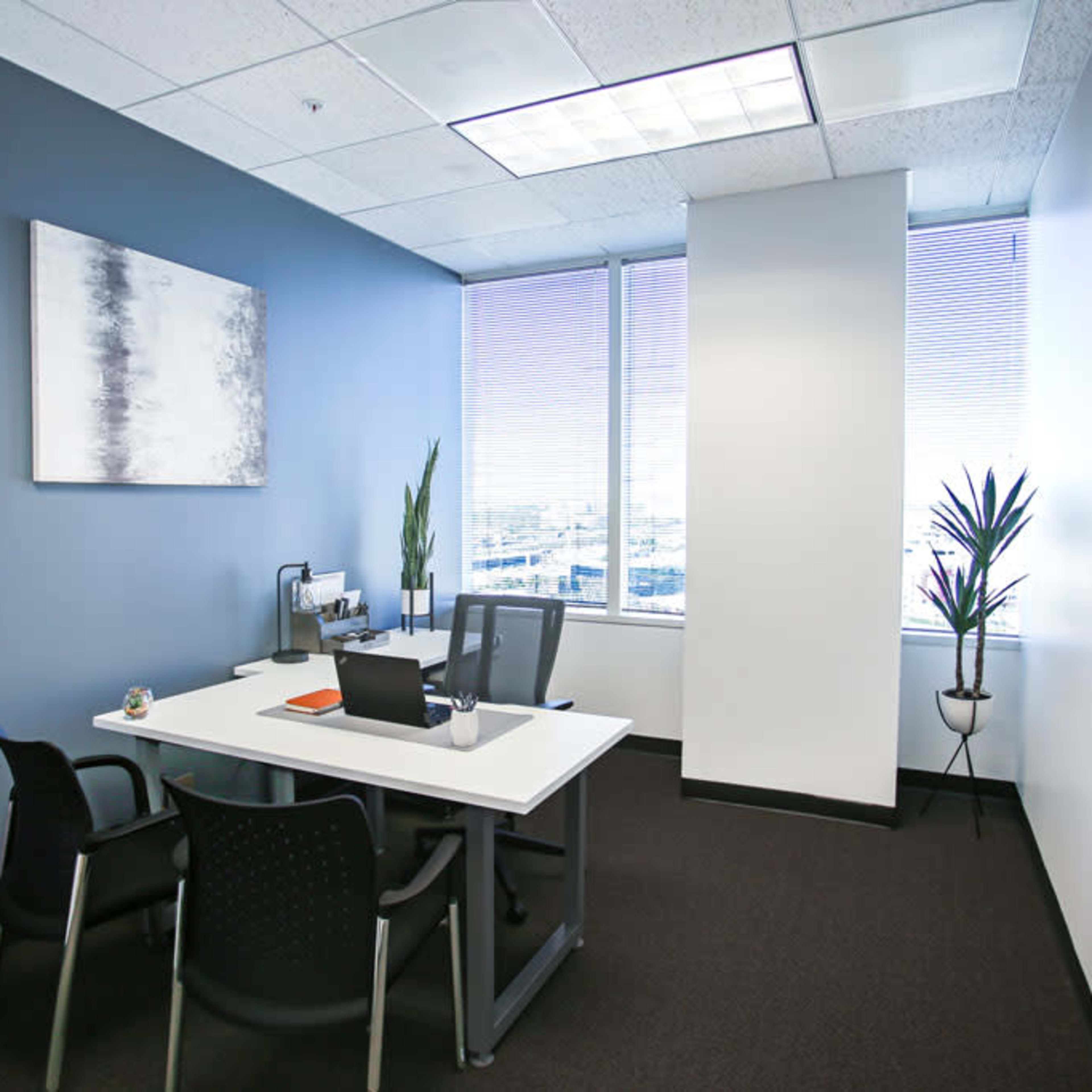 A small office space featuring a white desk with a computer, two chairs, and a potted plant beside a large window.