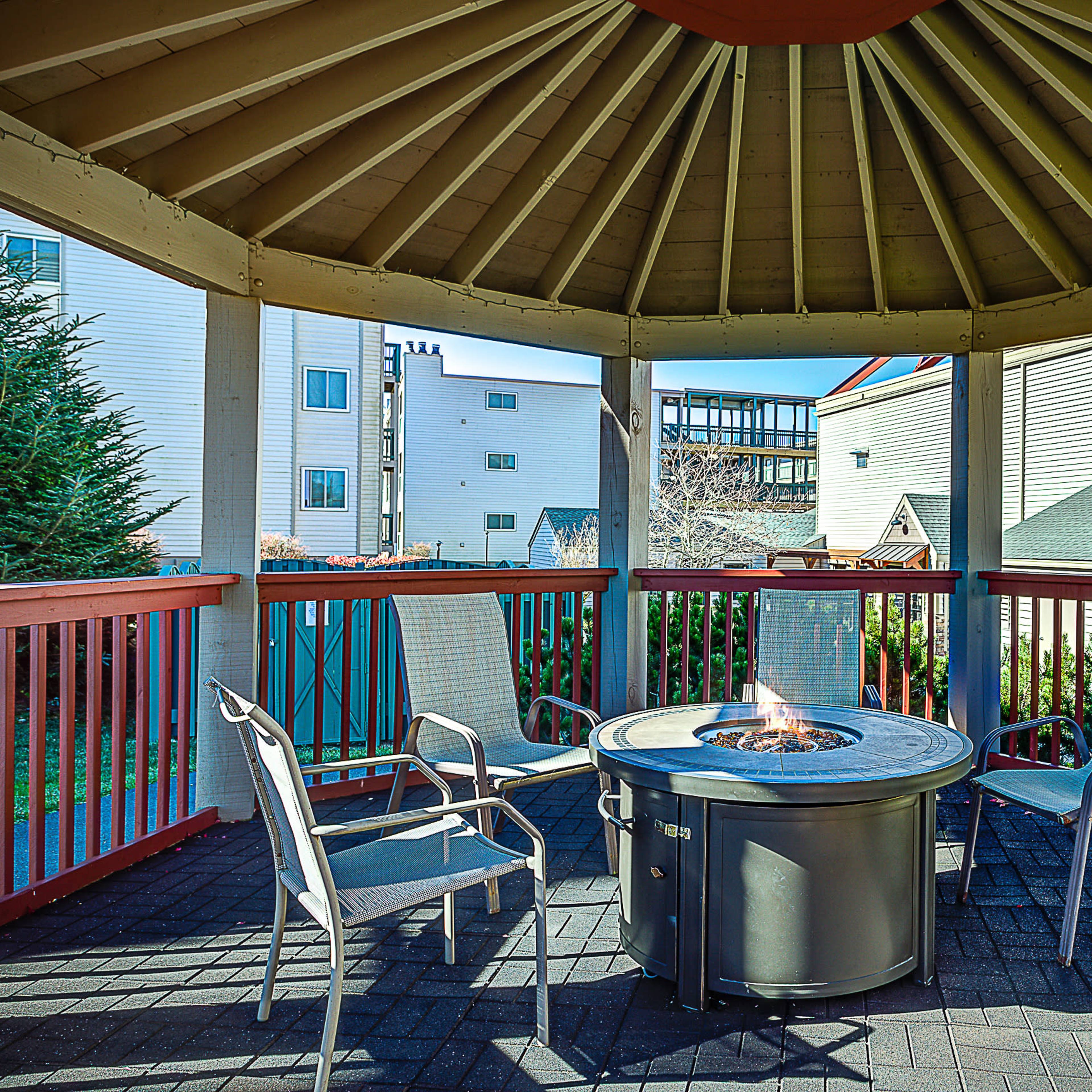 A gazebo with four metal chairs surrounding a circular fire pit on a stone patio.