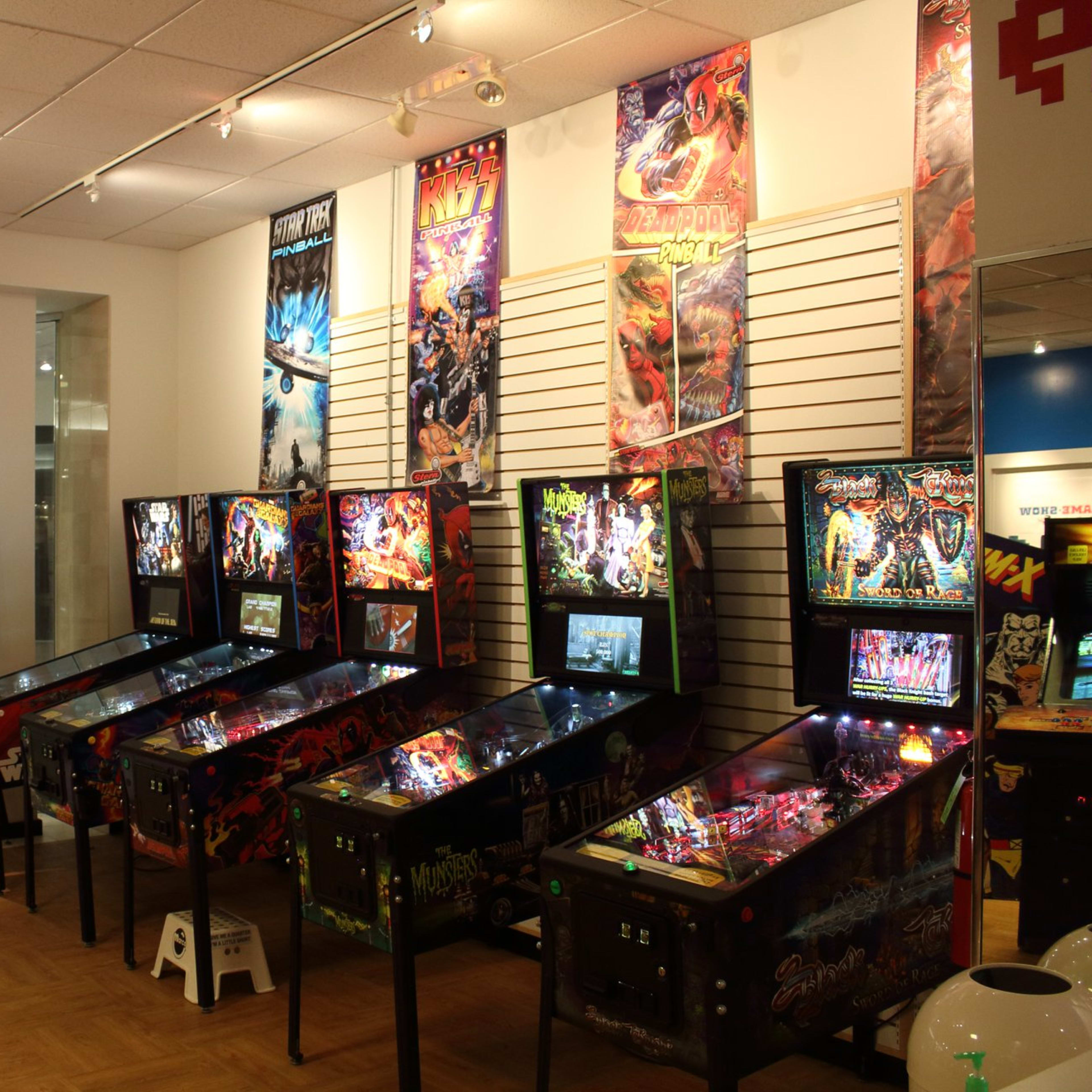 A row of pinball machines displayed in an arcade, with colorful posters hanging on the walls above them.