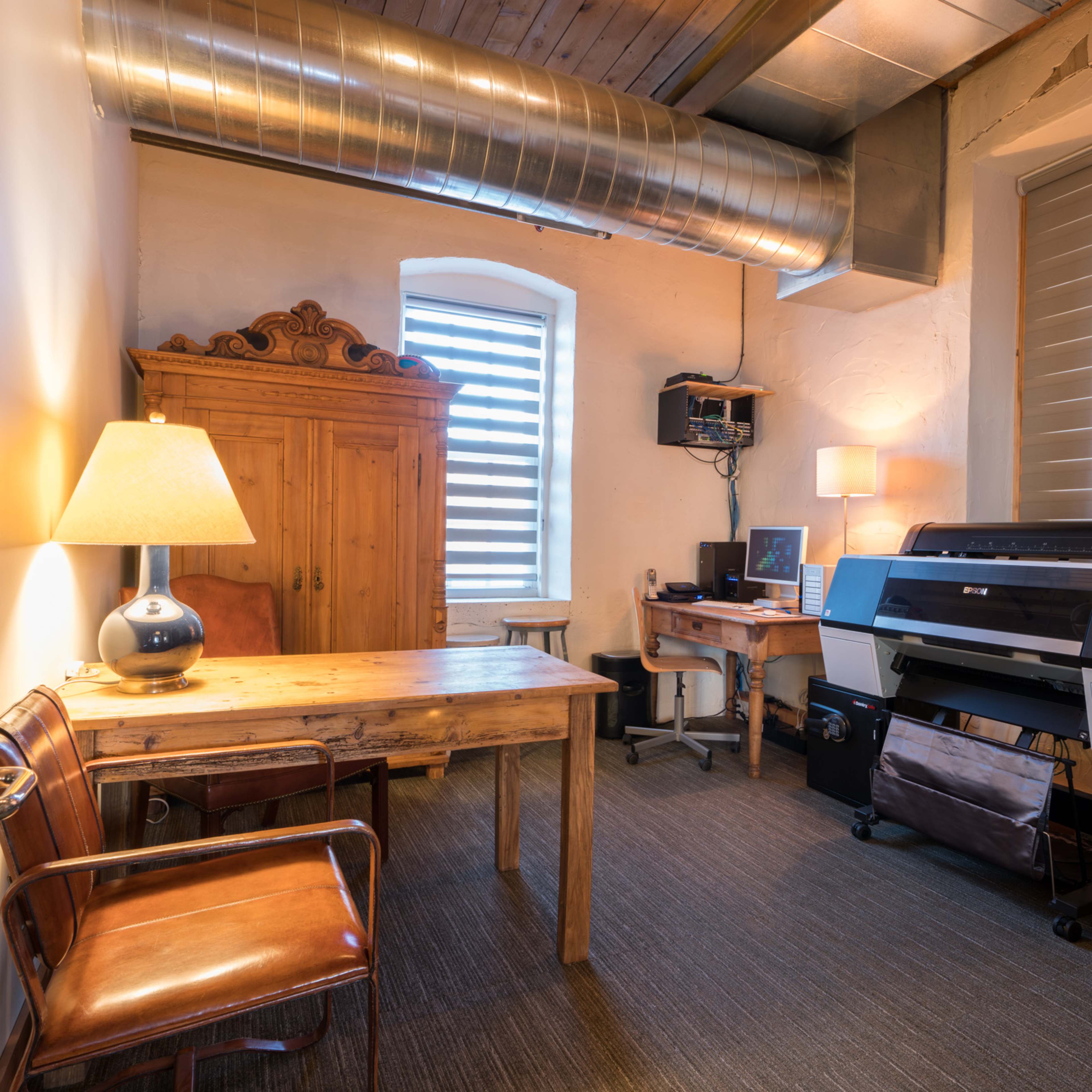 A small home office with a wooden table, two leather chairs, a large printer, and a vintage cabinet, all under a ceiling with exposed ductwork.