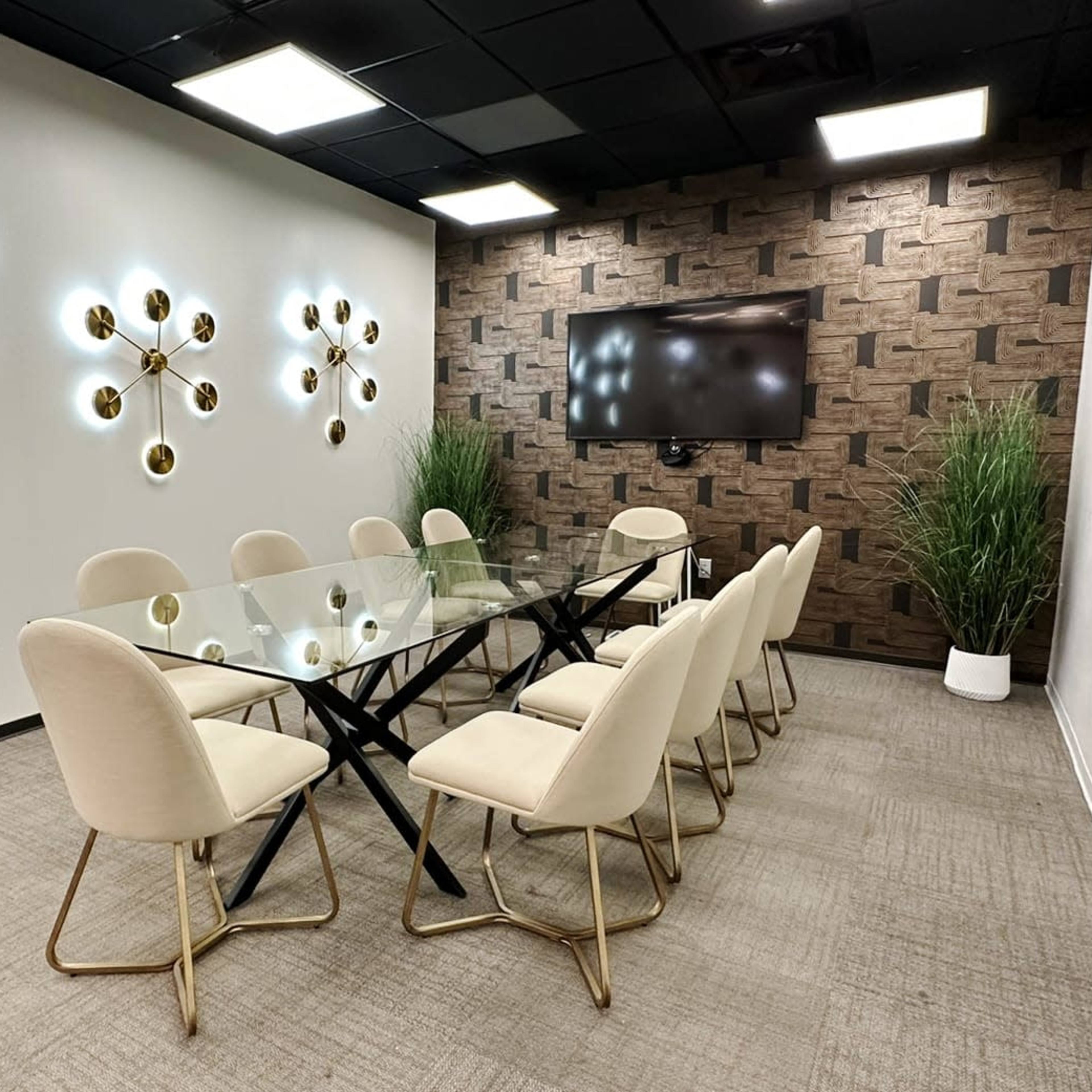 A modern conference room features a glass table surrounded by eight beige chairs, with stylish wall lights and a large screen mounted on a textured wall.
