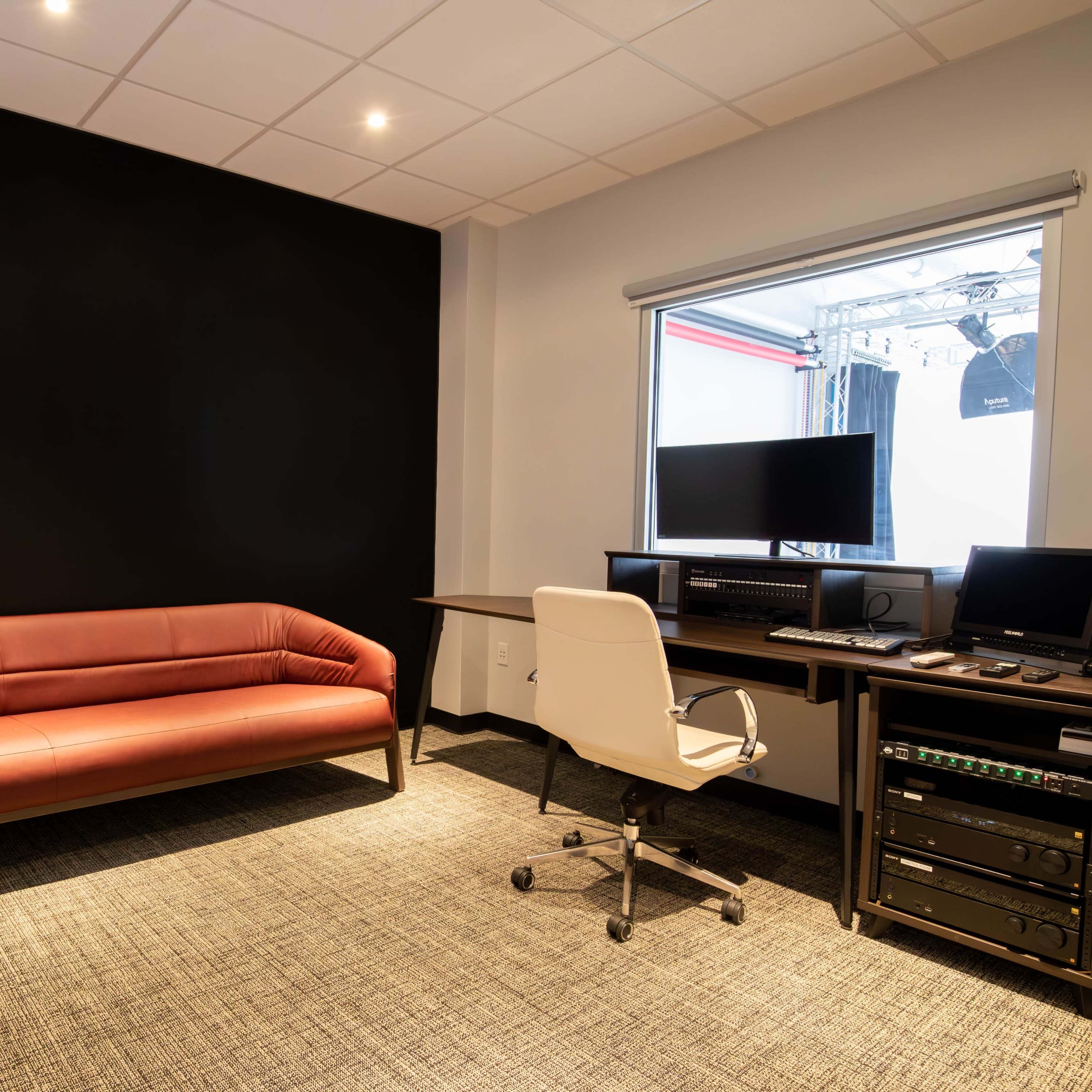 A modern office space with a brown sofa, a desk with computer equipment, and a large window allowing natural light.