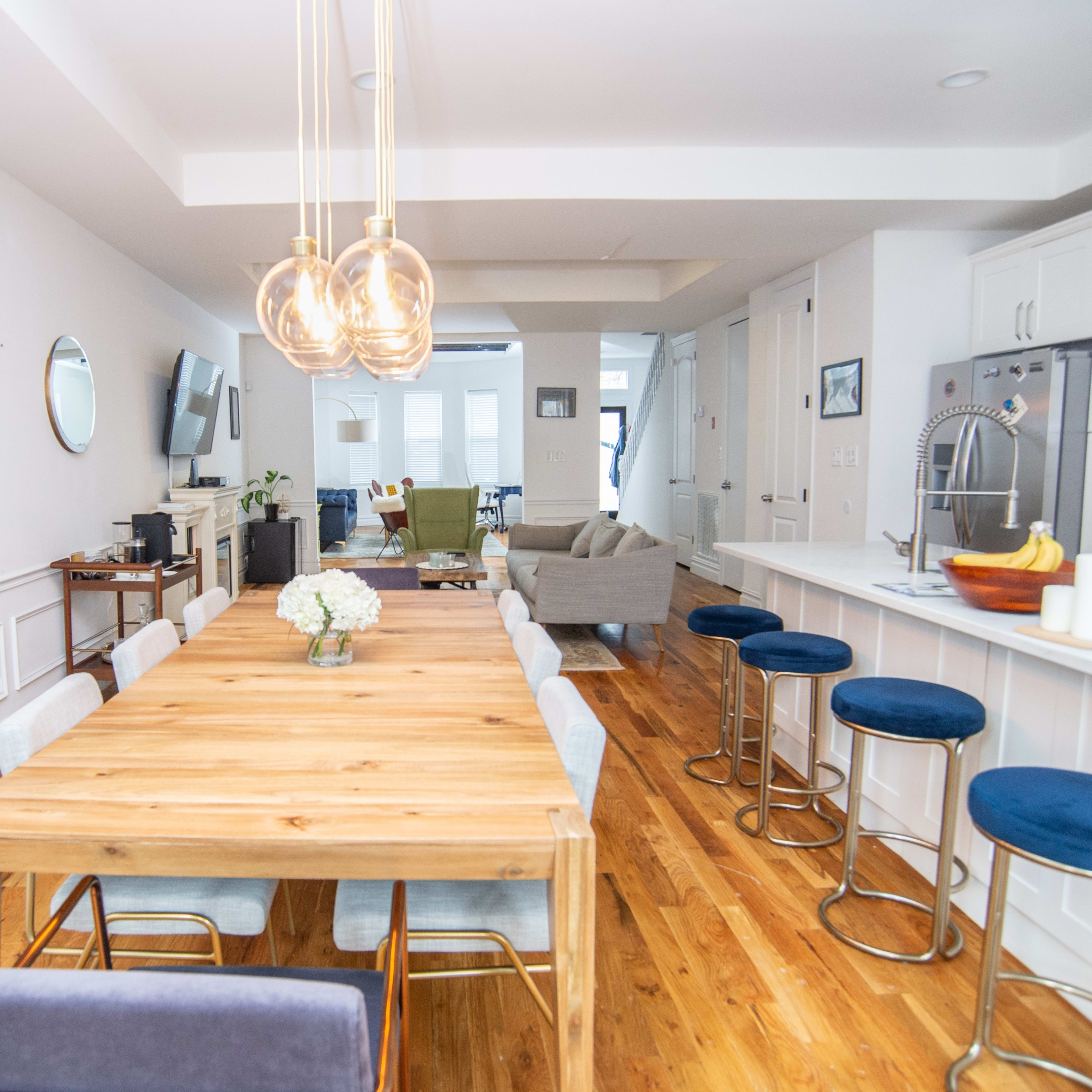 A modern dining area with a large wooden table surrounded by upholstered chairs, leading into a bright living space featuring a sofa and television.