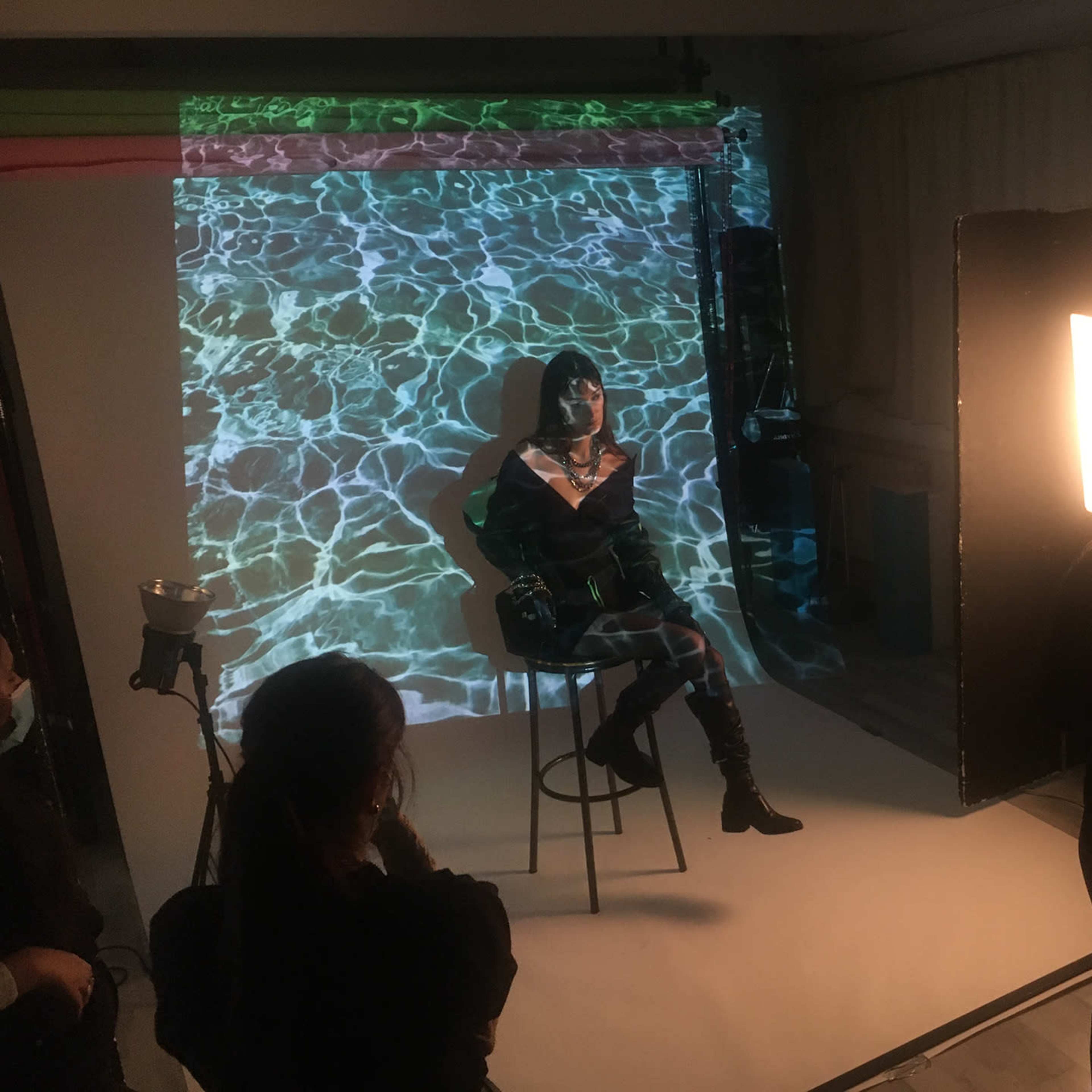 A model sits on a stool in front of a backdrop featuring a water pattern while being lit by a bright light during a photo shoot.