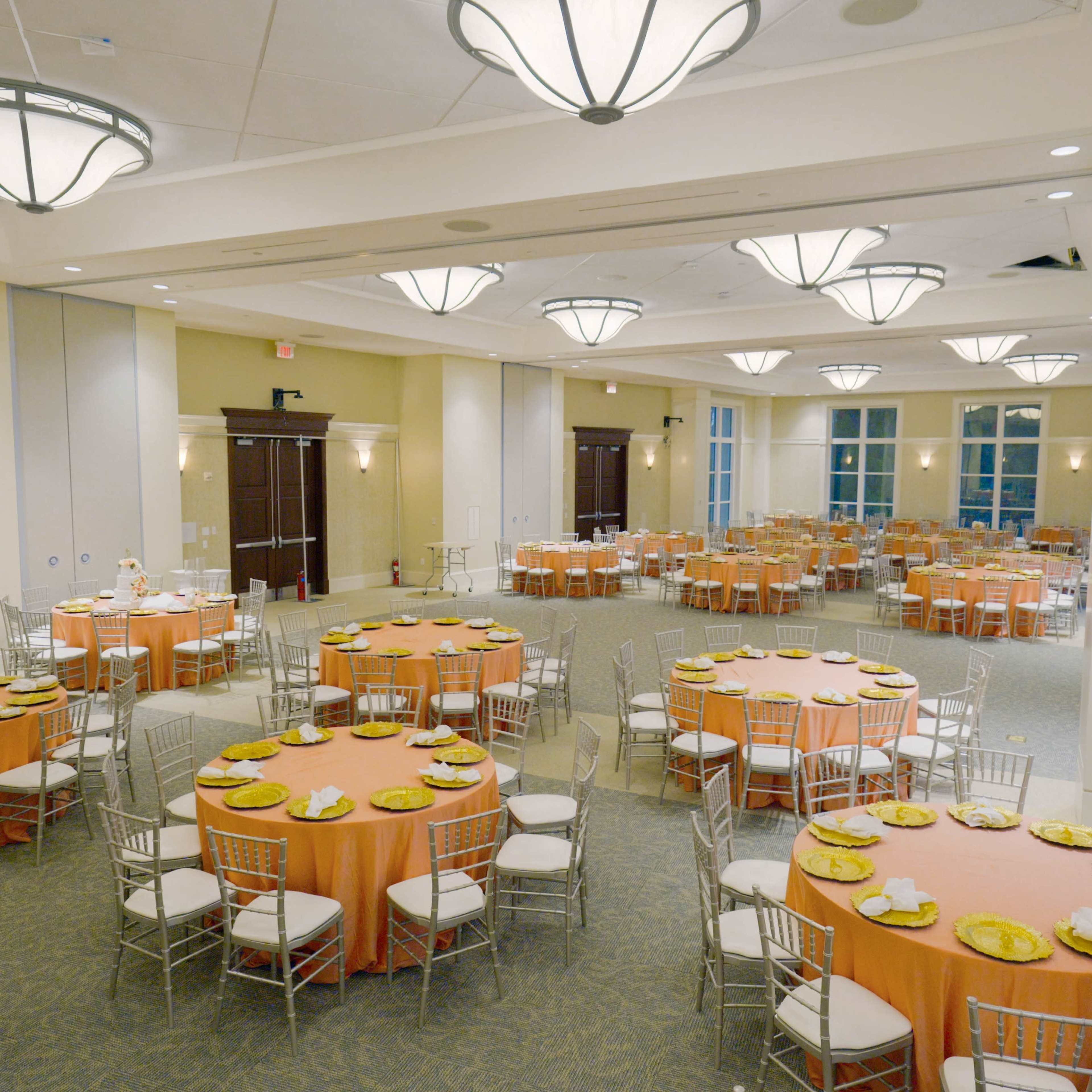 A banquet hall set with several round tables covered in orange tablecloths, each adorned with plates and silverware, under modern ceiling lights.